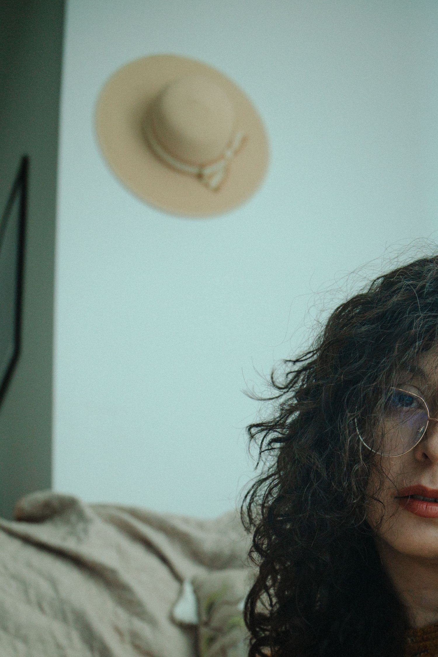 A figure with curly dark hair and red lipstick wearing a beige hat is shown in an artistic profile shot.