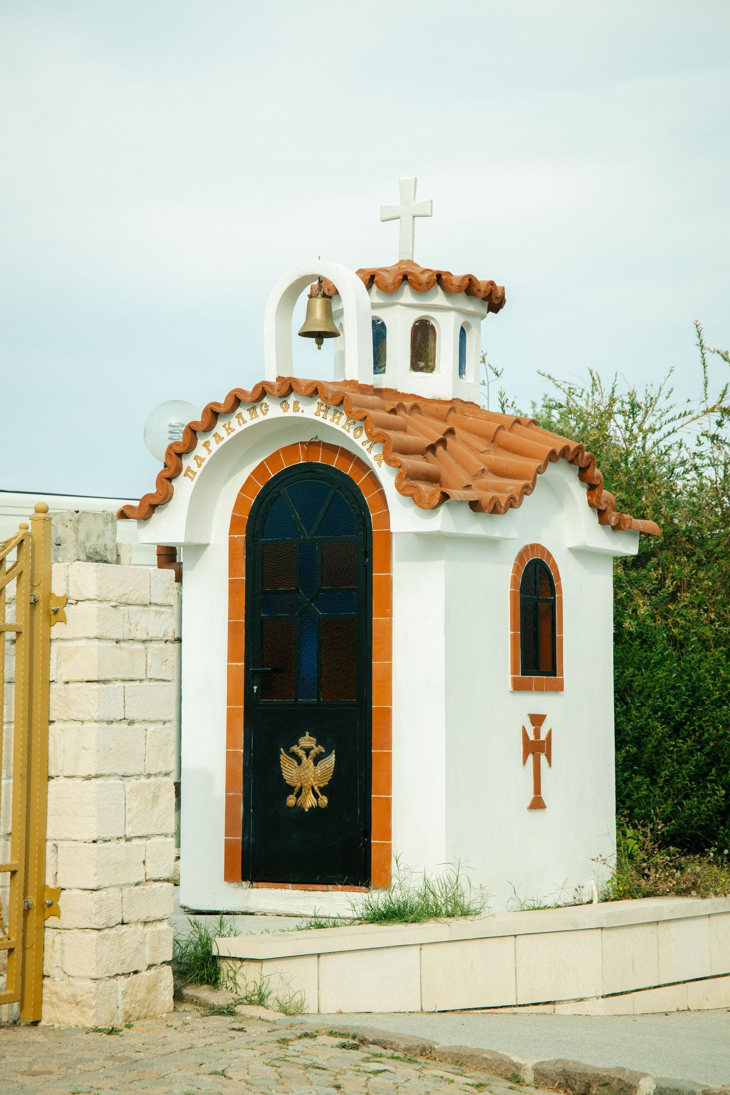 Small white Greek Orthodox roadside chapel with terracotta roof, bell, cross, and ornate double-headed eagle door symbol.