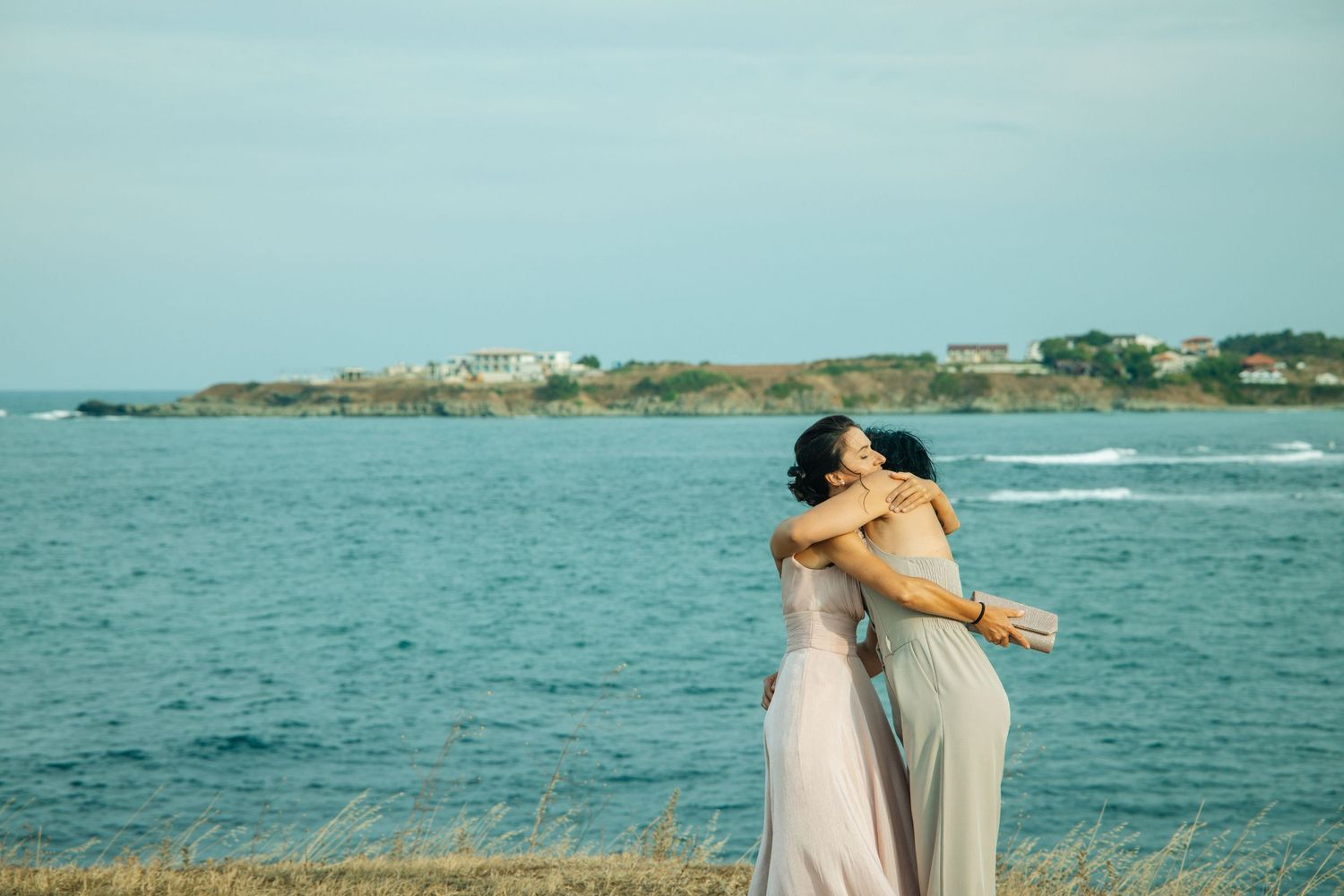 Two women in elegant gowns embrace near rocky ocean coastline under a clear blue sky.