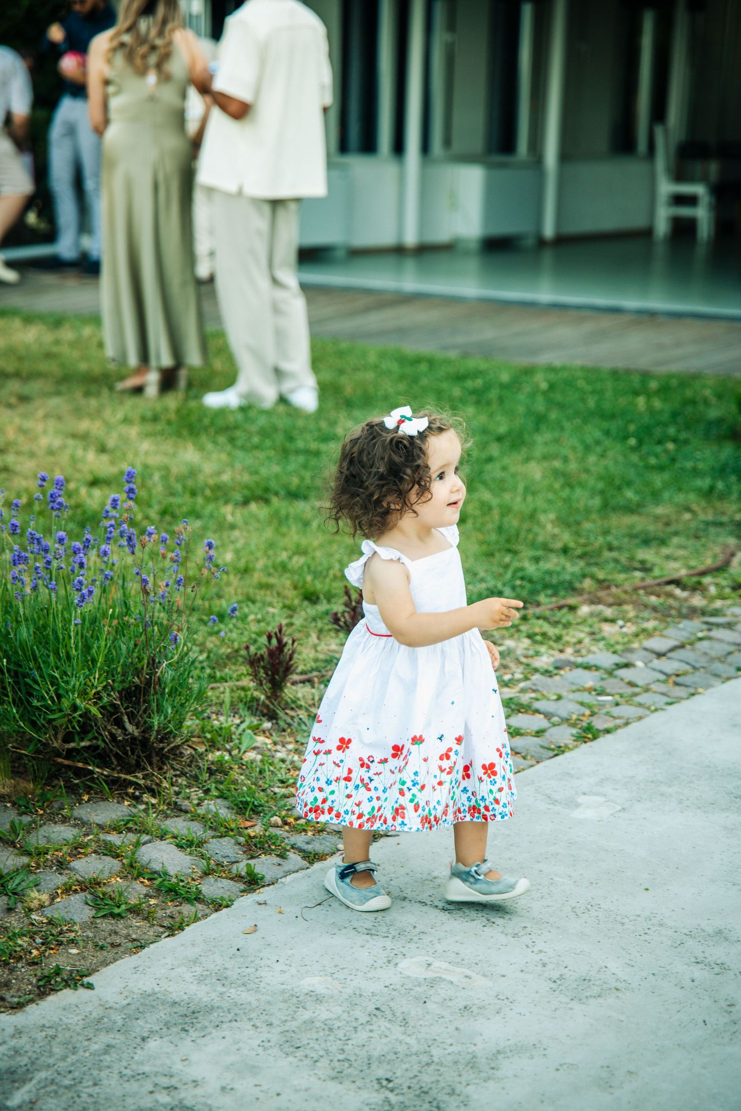 Toddler girl in white floral dress standing on garden path at outdoor wedding.