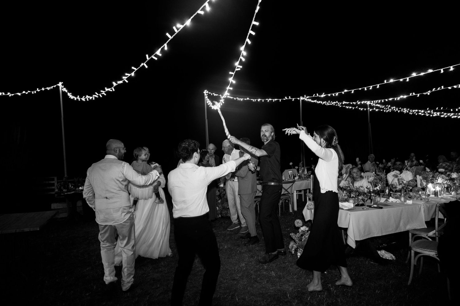 Guests dance under string lights at an outdoor wedding reception in black and white.