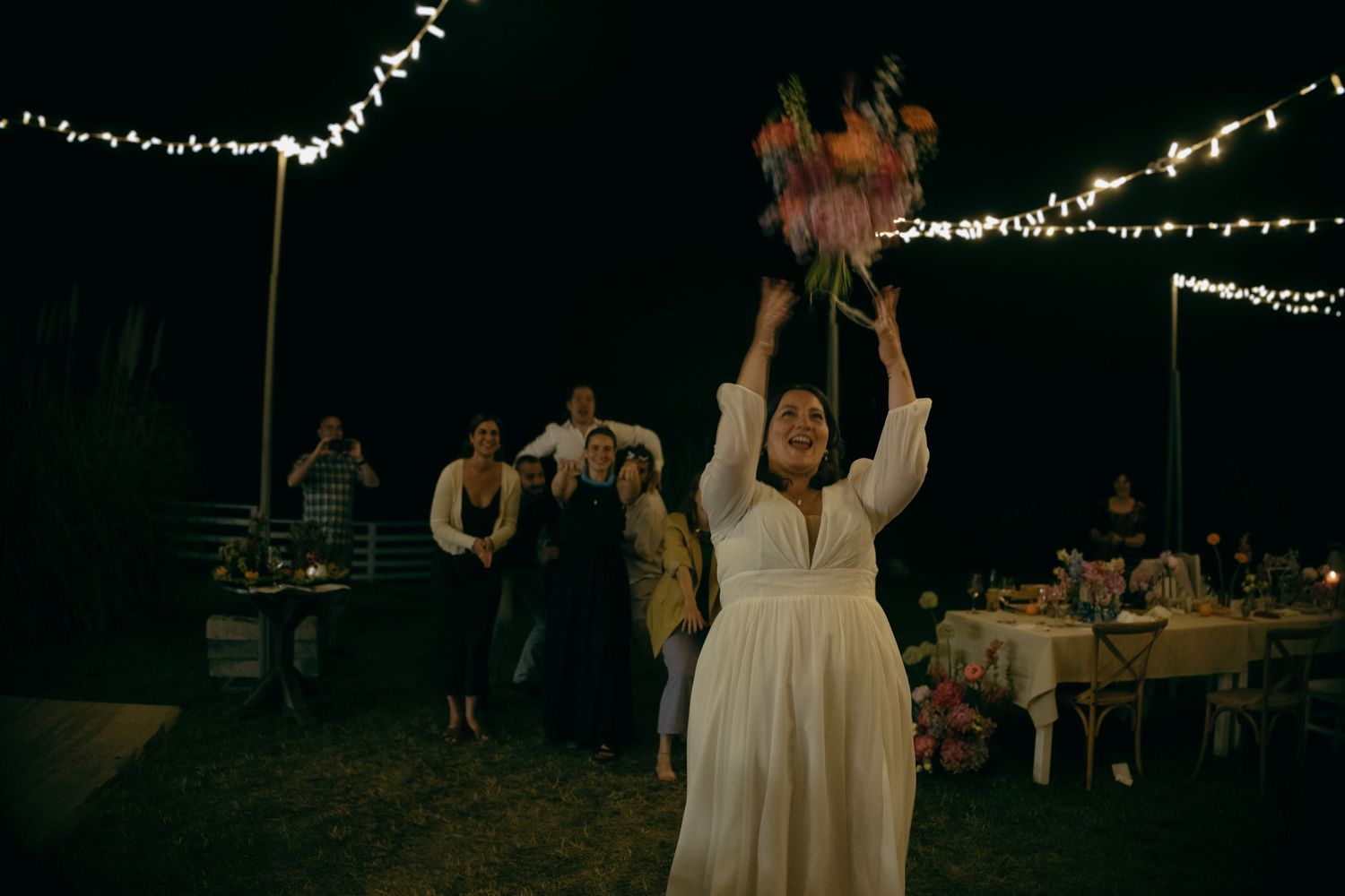 Bride tossing bouquet at outdoor evening wedding reception with string lights and guests in background.