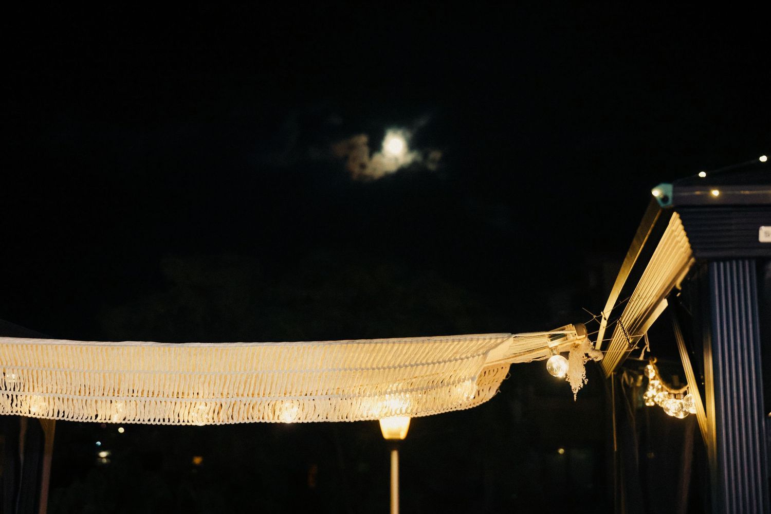 Moonlit night sky above a decorated outdoor canopy with warm string lights and lace fabric.