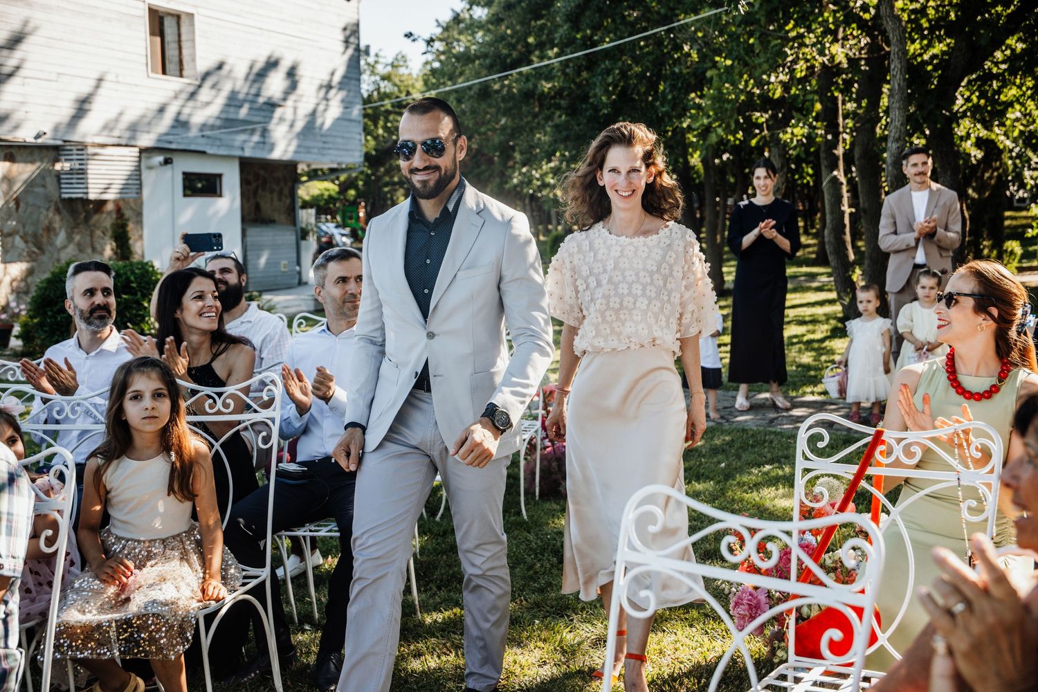 Couple walks down outdoor wedding aisle surrounded by seated guests on a sunny day.