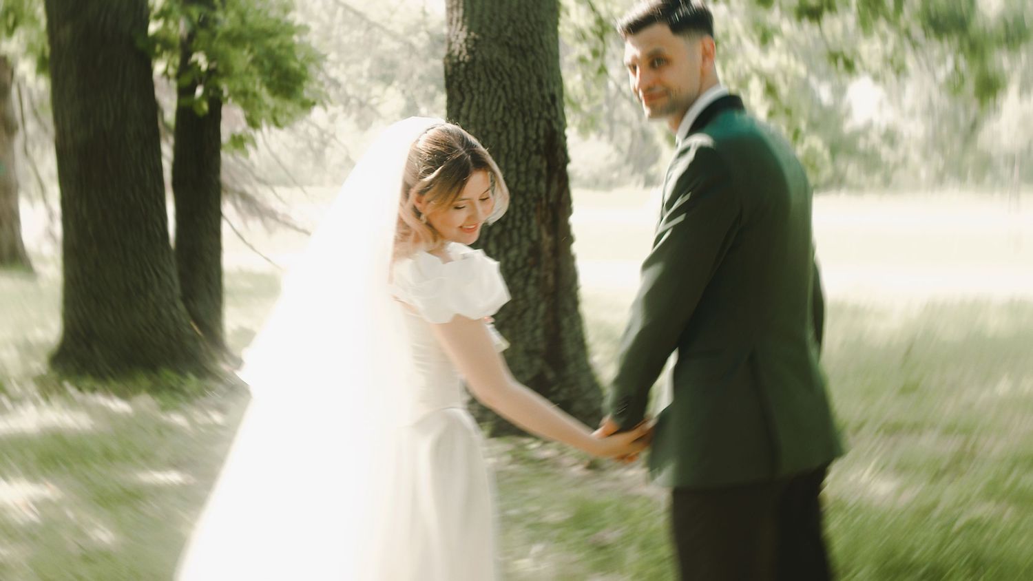 A bride in a white gown and veil holds hands with a groom in a green suit outdoors among lush green trees.