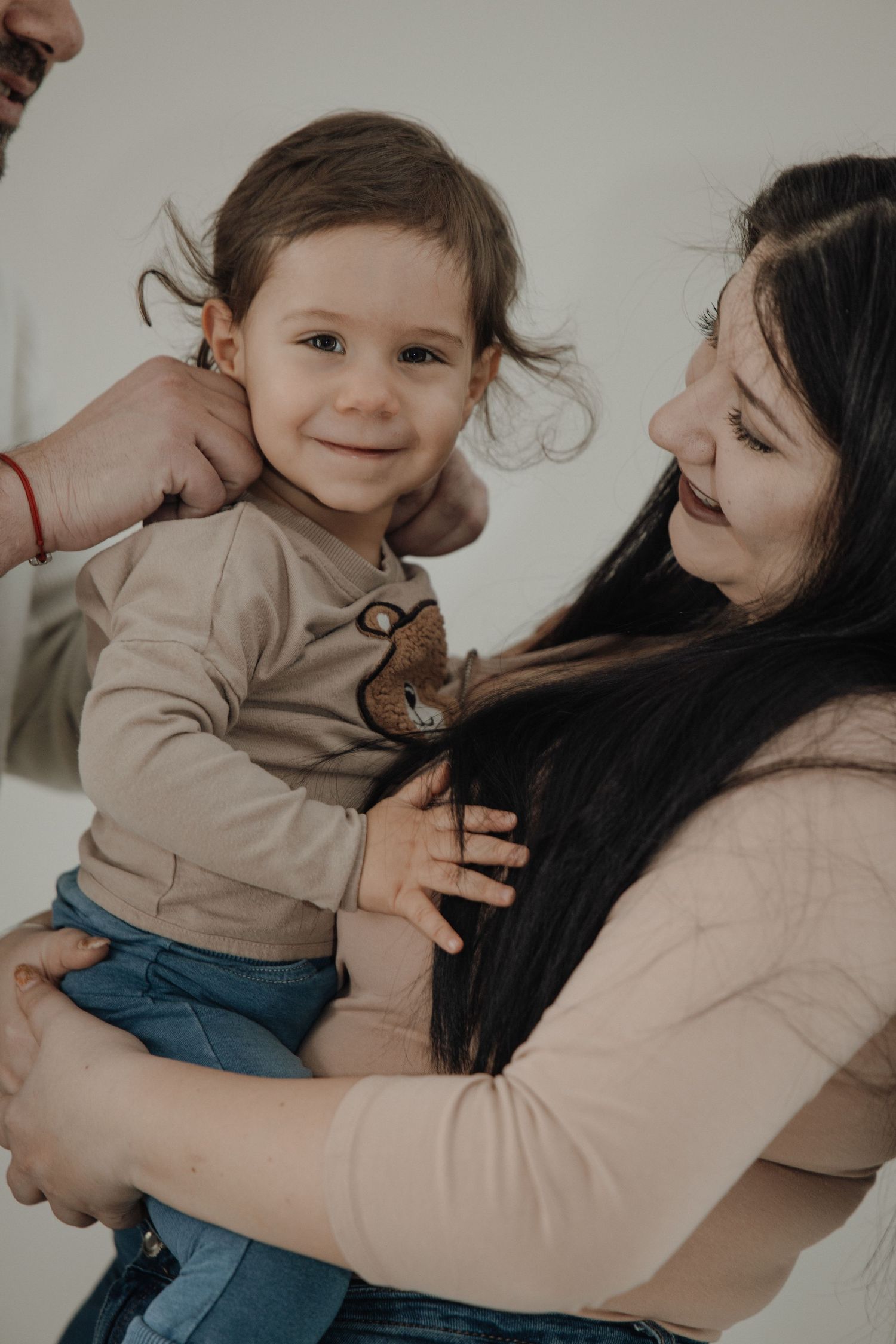 A smiling toddler being held by a dark-haired woman while another person touches the child's hair.