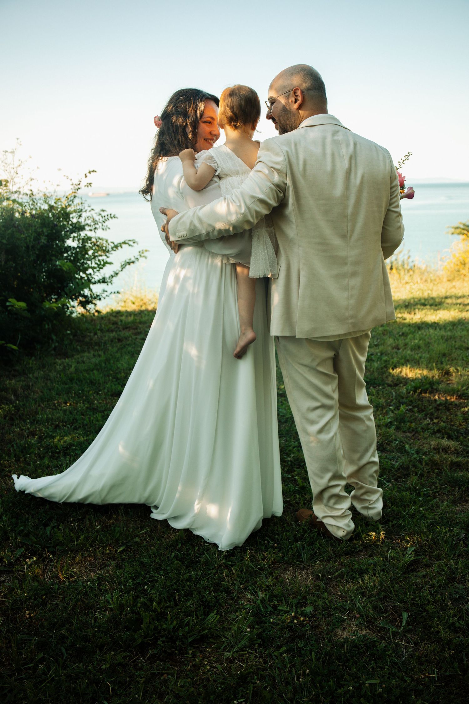 Bride and groom in white and beige wedding attire holding baby by seaside at sunset.