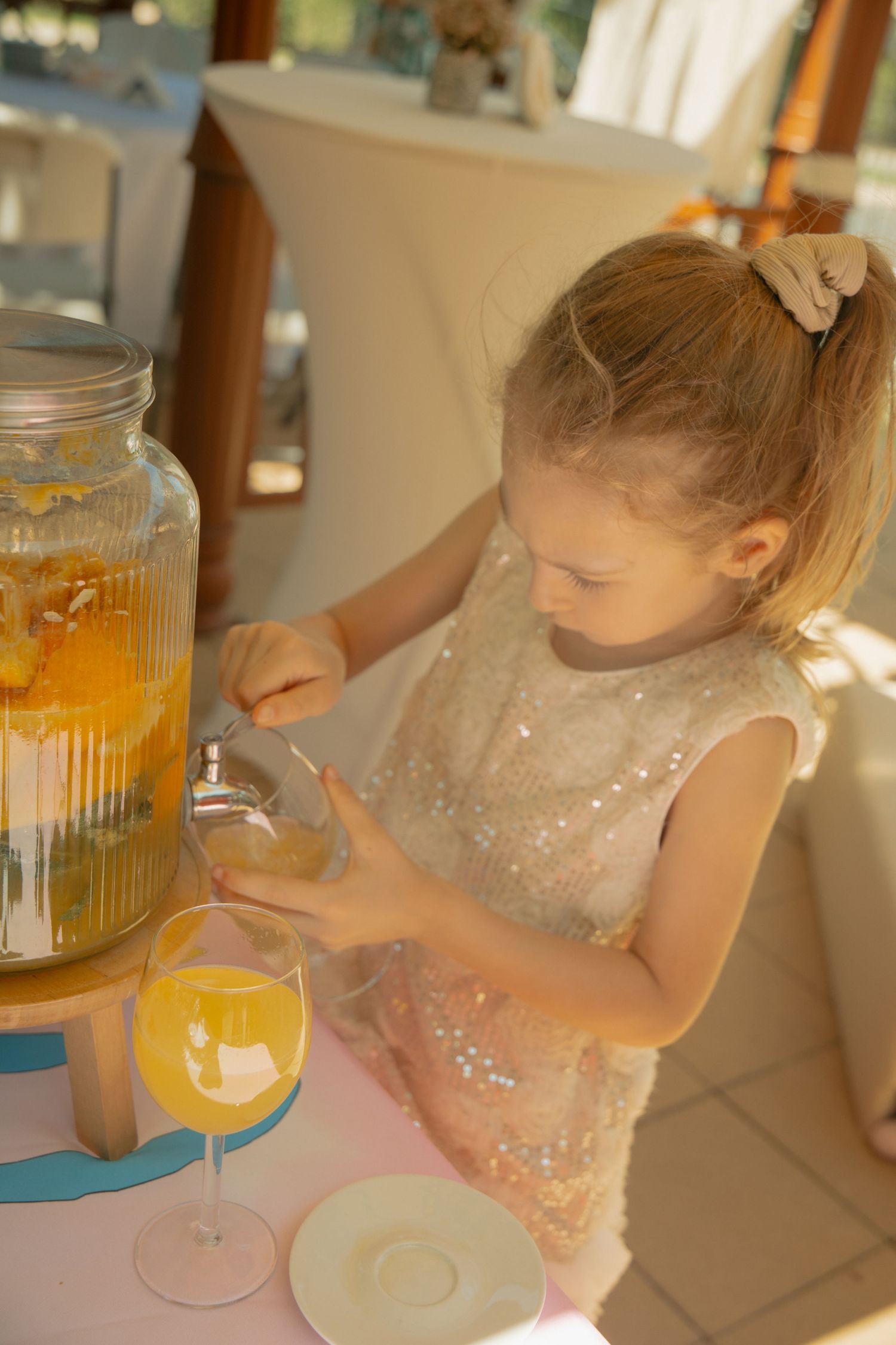 Young girl in sequin dress pouring orange juice into a wine glass at a decorated table.