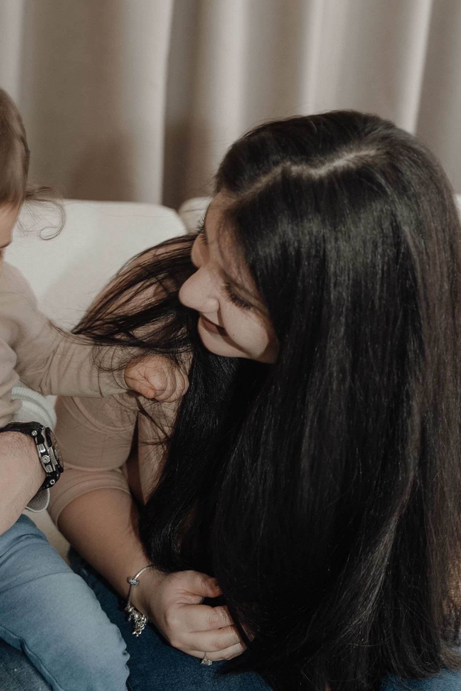 A smiling woman with long dark hair plays with a young child indoors, wearing a smartwatch and bracelet.
