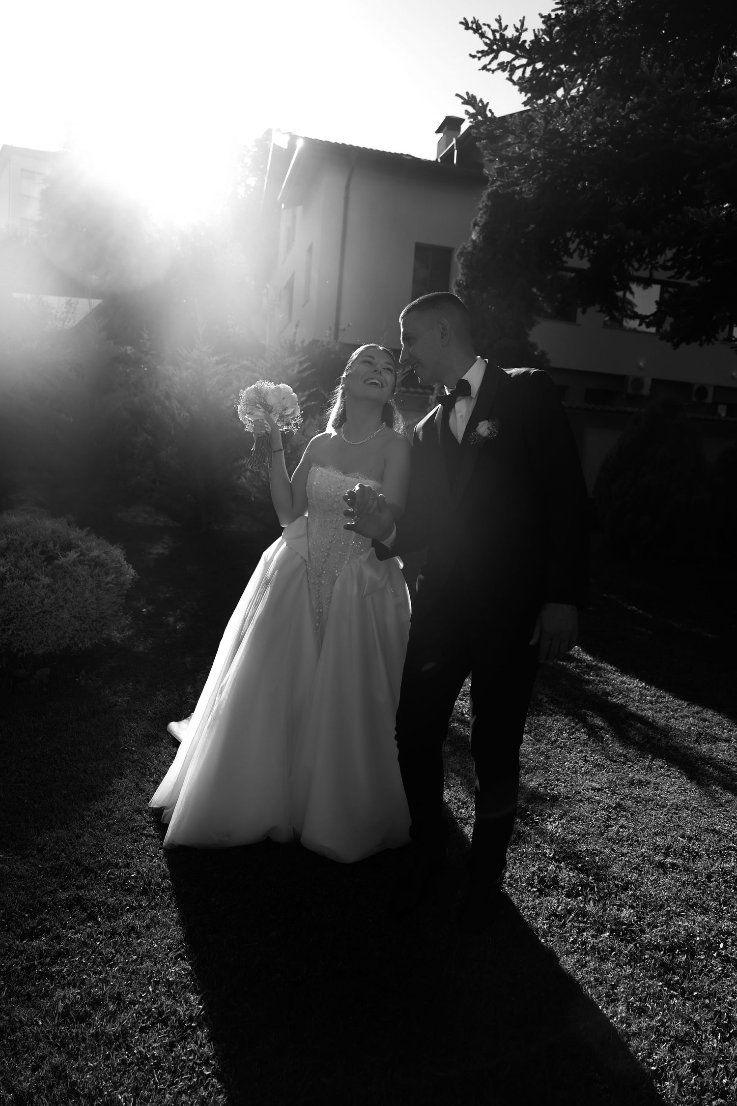 Black and white wedding photo of bride and groom embracing outdoors with dramatic sunlight behind them.