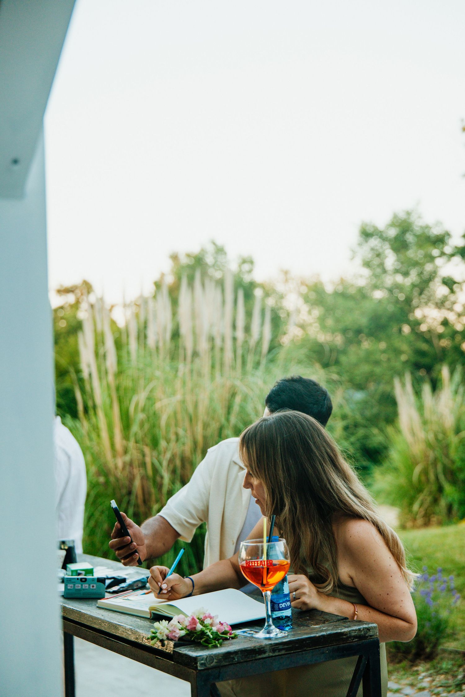 Woman signing documents at outdoor wedding table with Aperol spritz and flowers, lush greenery backdrop.