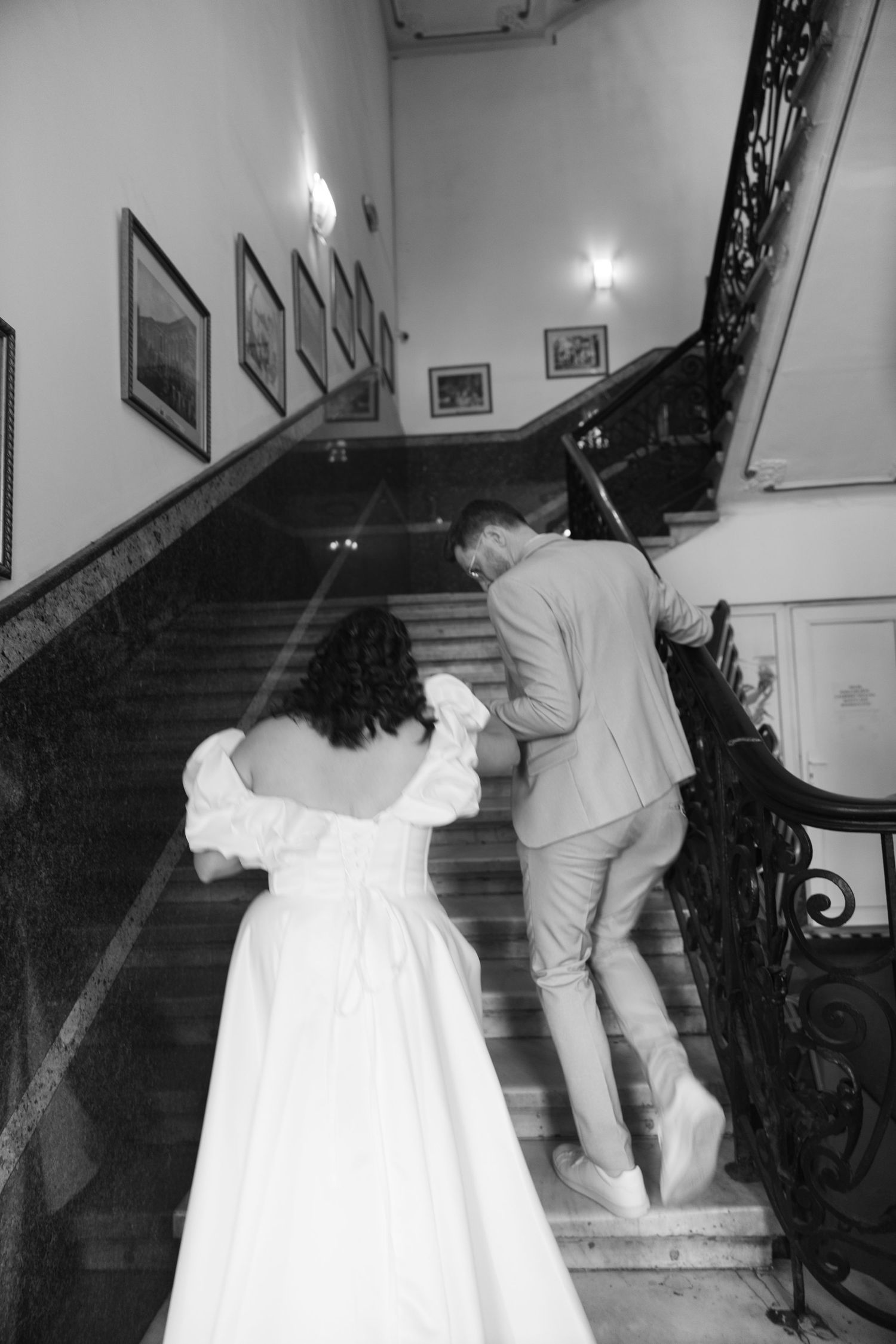 Black and white photo of a bride and groom walking up an elegant staircase, holding hands at their wedding venue.