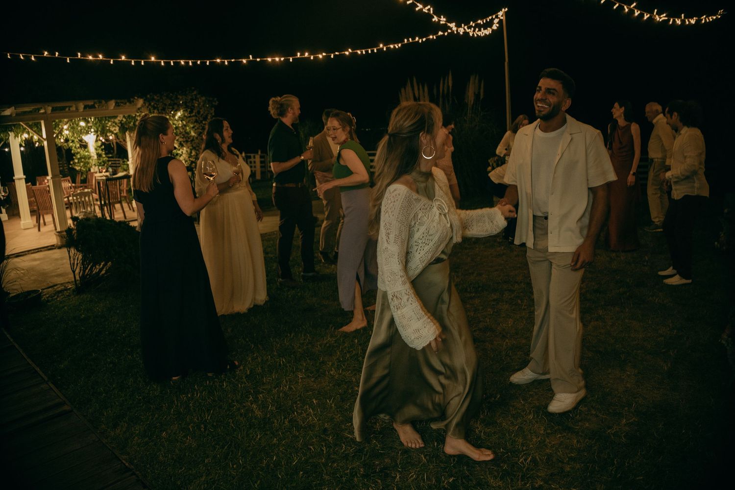 Guests dance and laugh together on a lawn at night under string lights at an outdoor party.