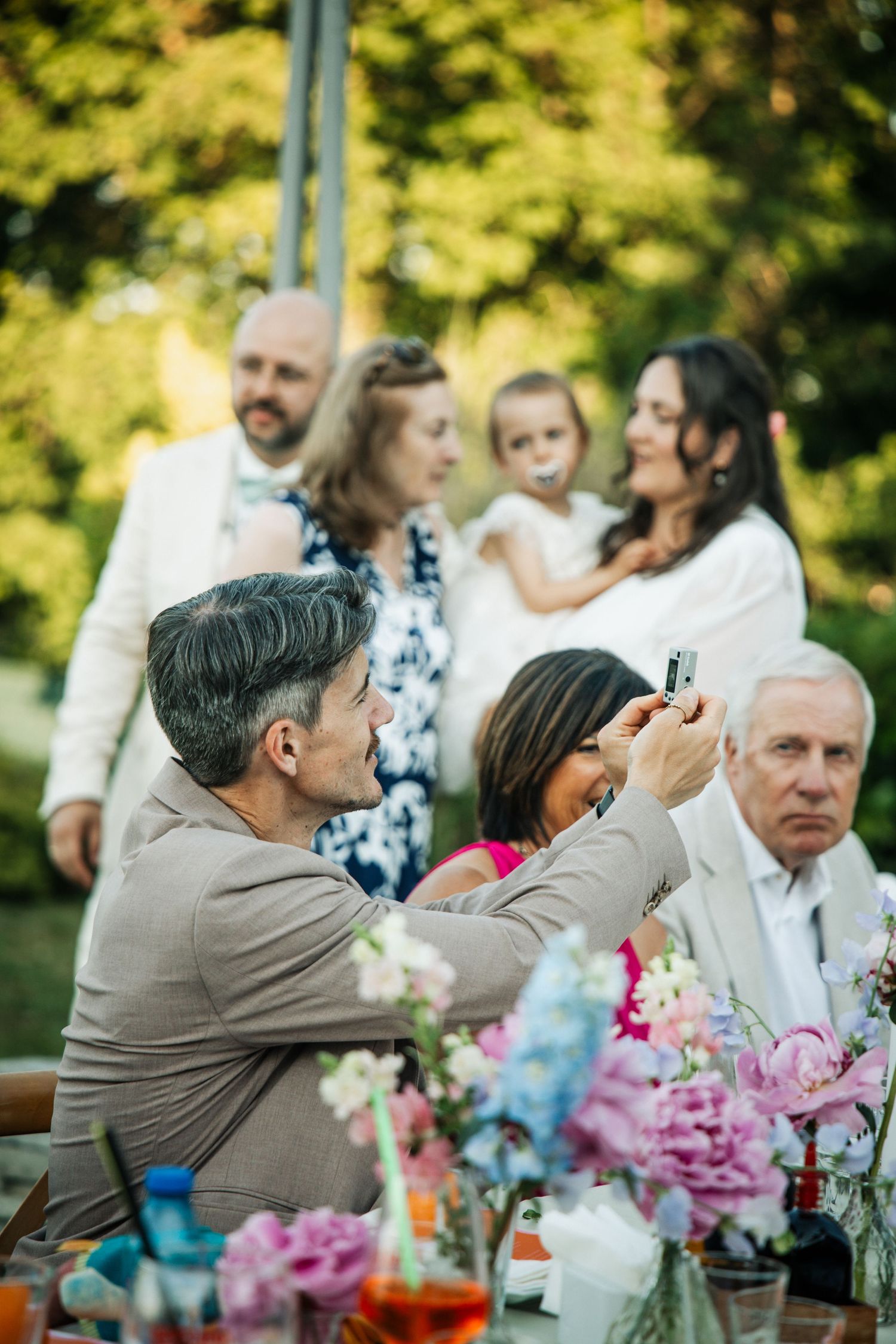 Guests seated at an outdoor wedding reception table with flowers, while a man photographs a family posing behind.