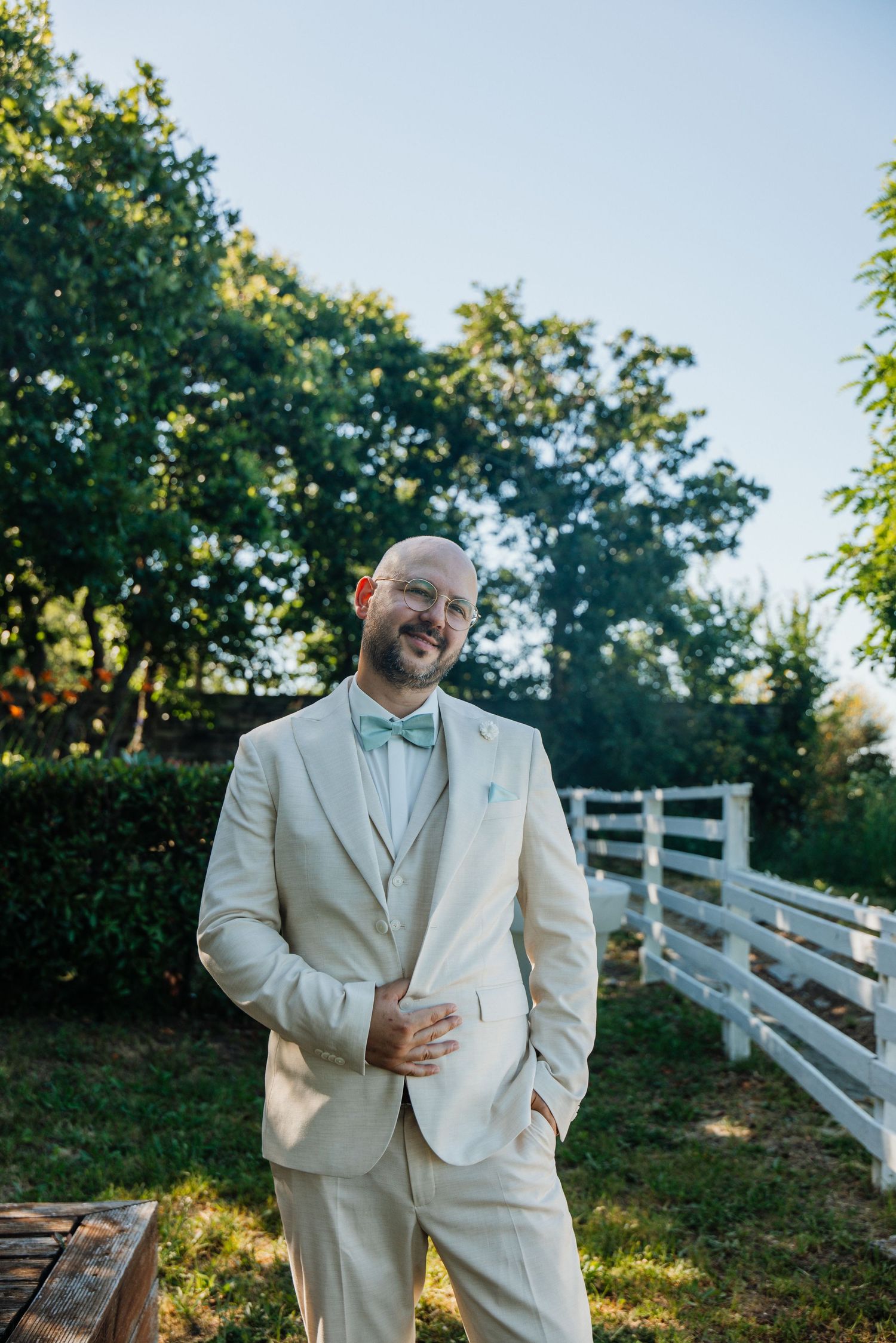 Smiling groom in beige suit with mint bow tie stands outdoors near white fence.