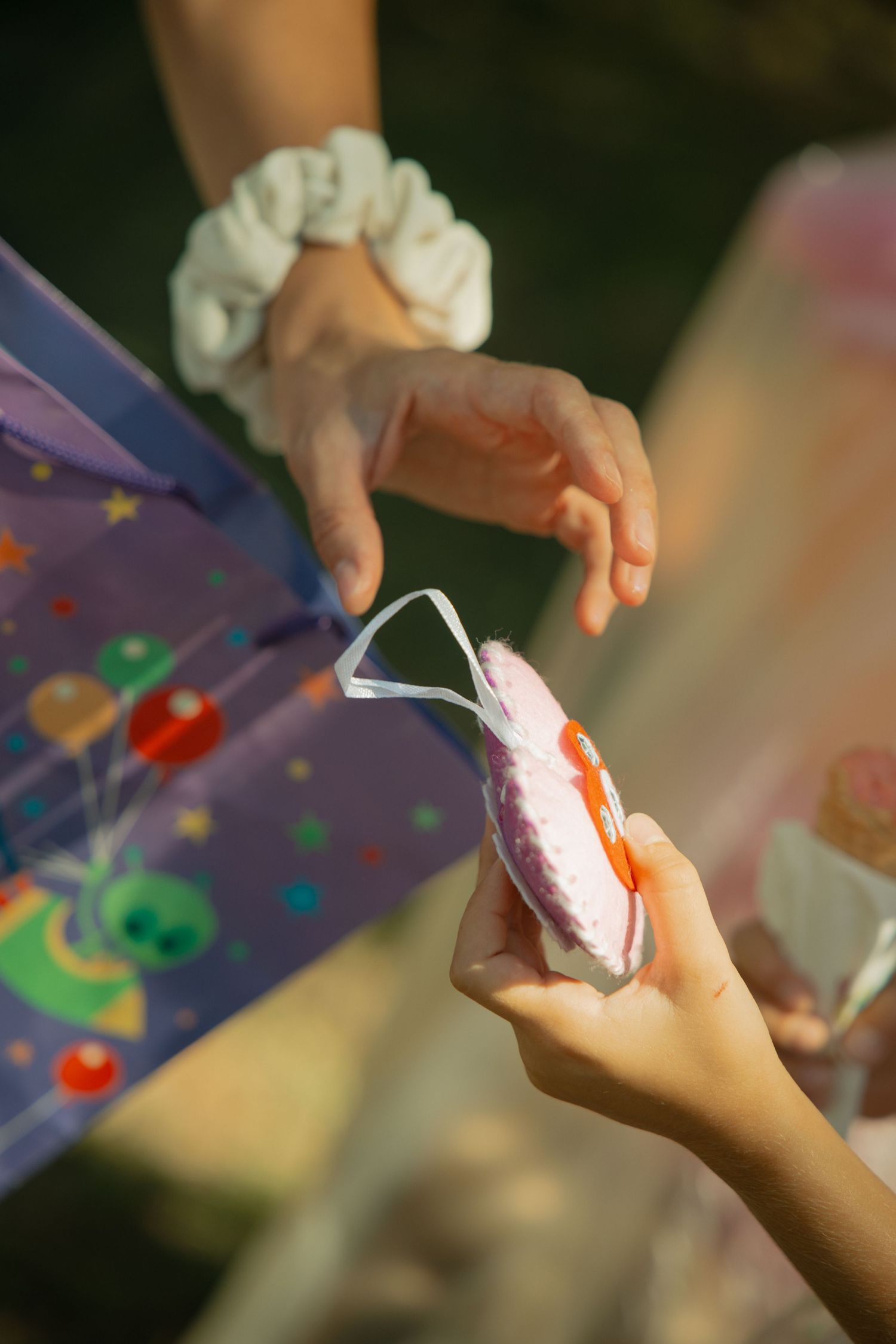 Child receiving a pink macaron treat from an adult wearing a white scrunchie at a colorful birthday party.