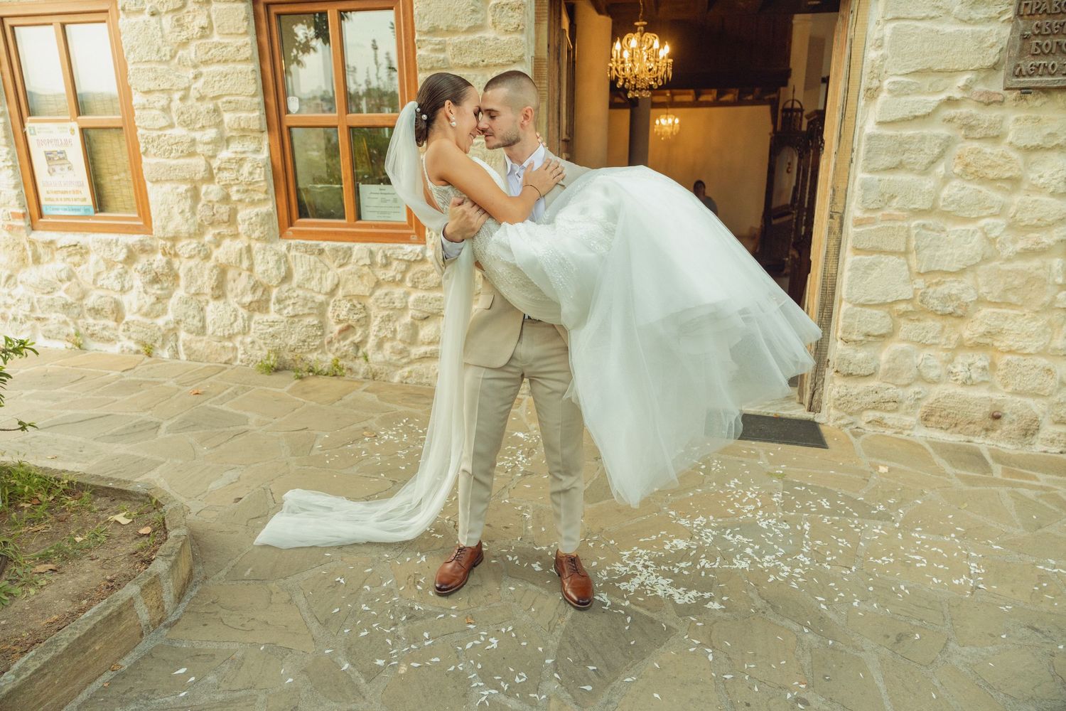Groom lifts bride in white wedding dress outside stone building on their wedding day.