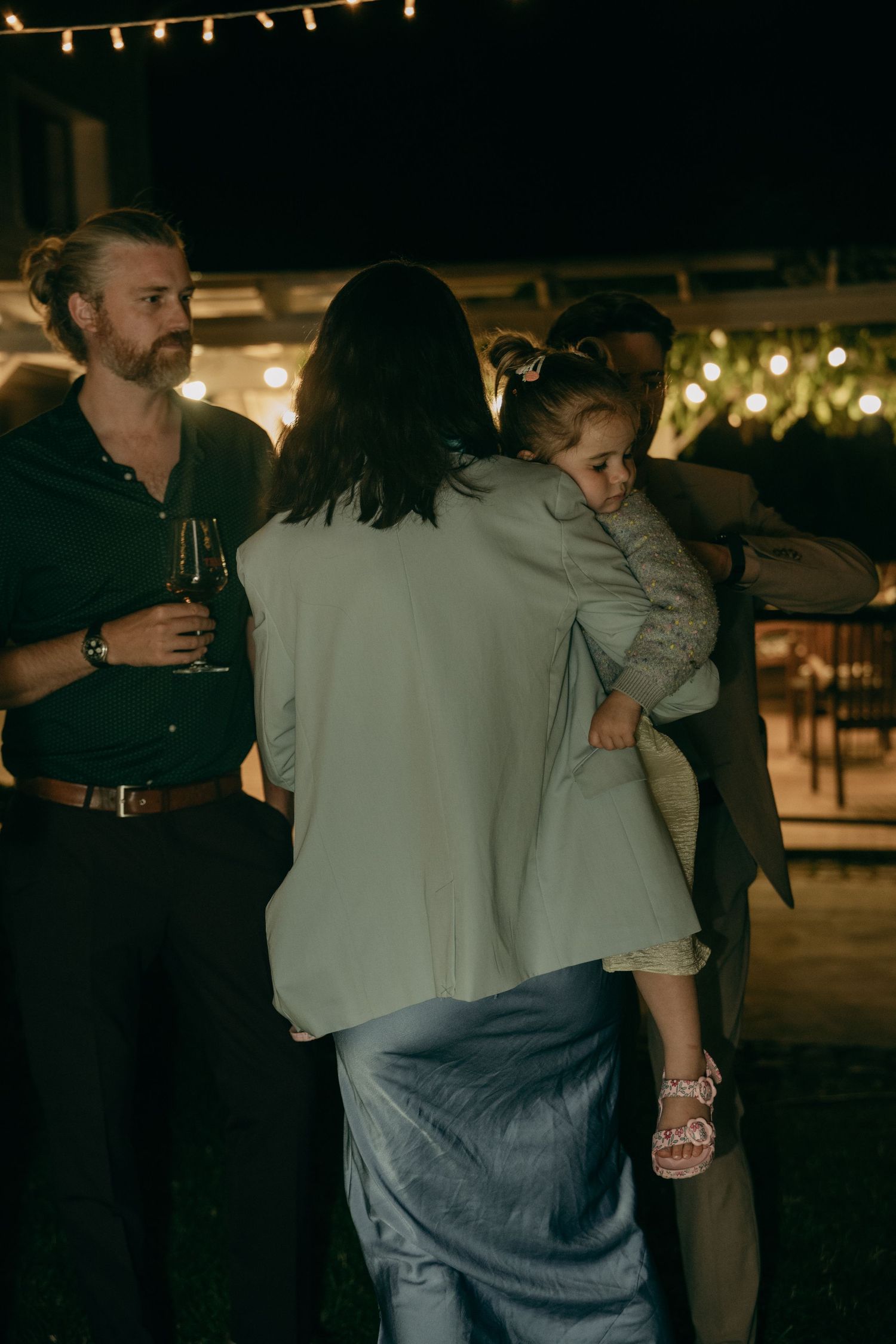 Adults and a child gather at an outdoor evening party with string lights, one man holding a wine glass.