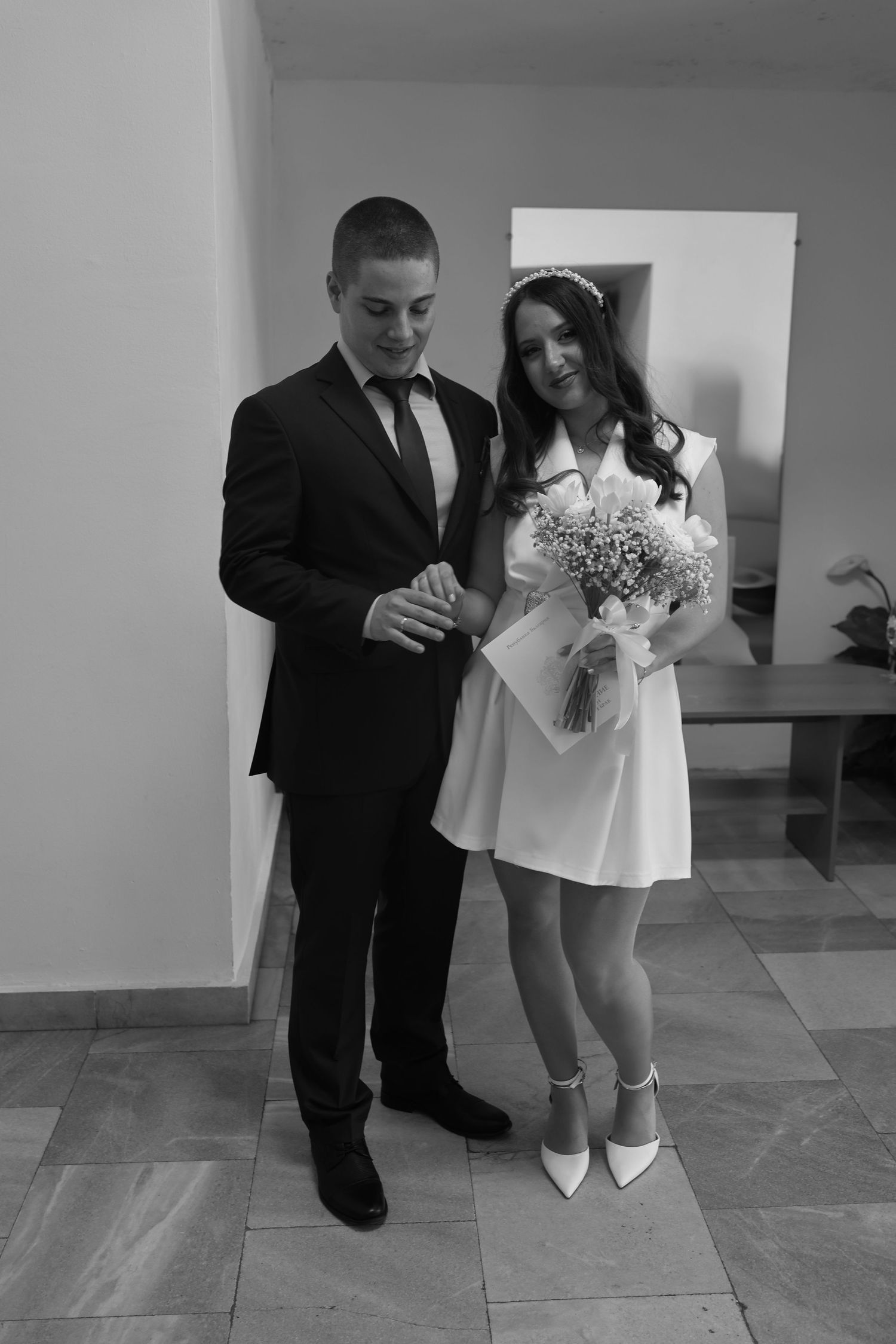 Black and white photo of a young couple in wedding attire, groom in suit and bride in short white dress holding a bouquet.