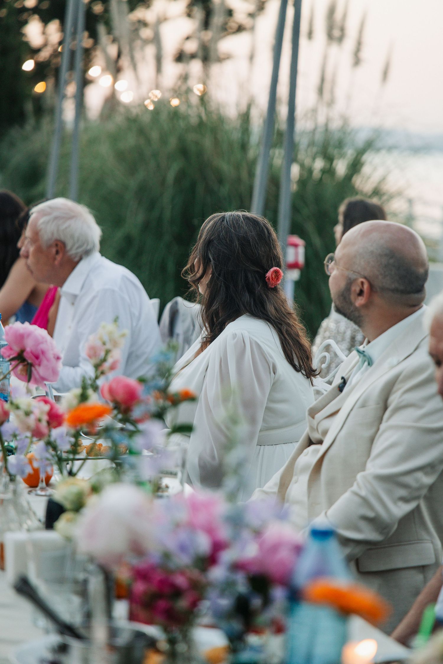 Elegant outdoor wedding reception at dusk with colorful floral centerpieces and guests in white attire seated by the water.