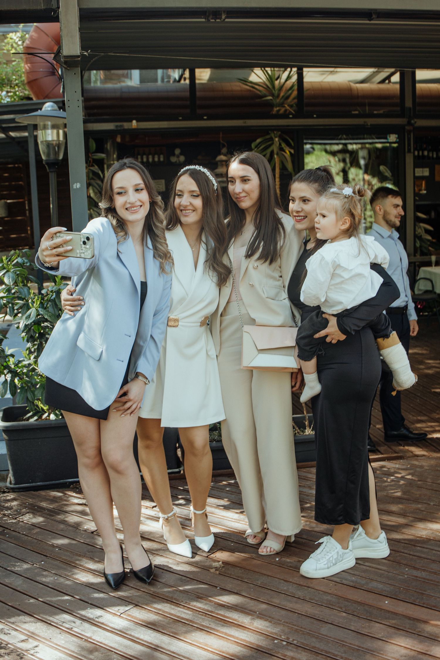 Four women posing for a selfie outdoors at an event, one holding a child, dressed in elegant white and blue outfits.