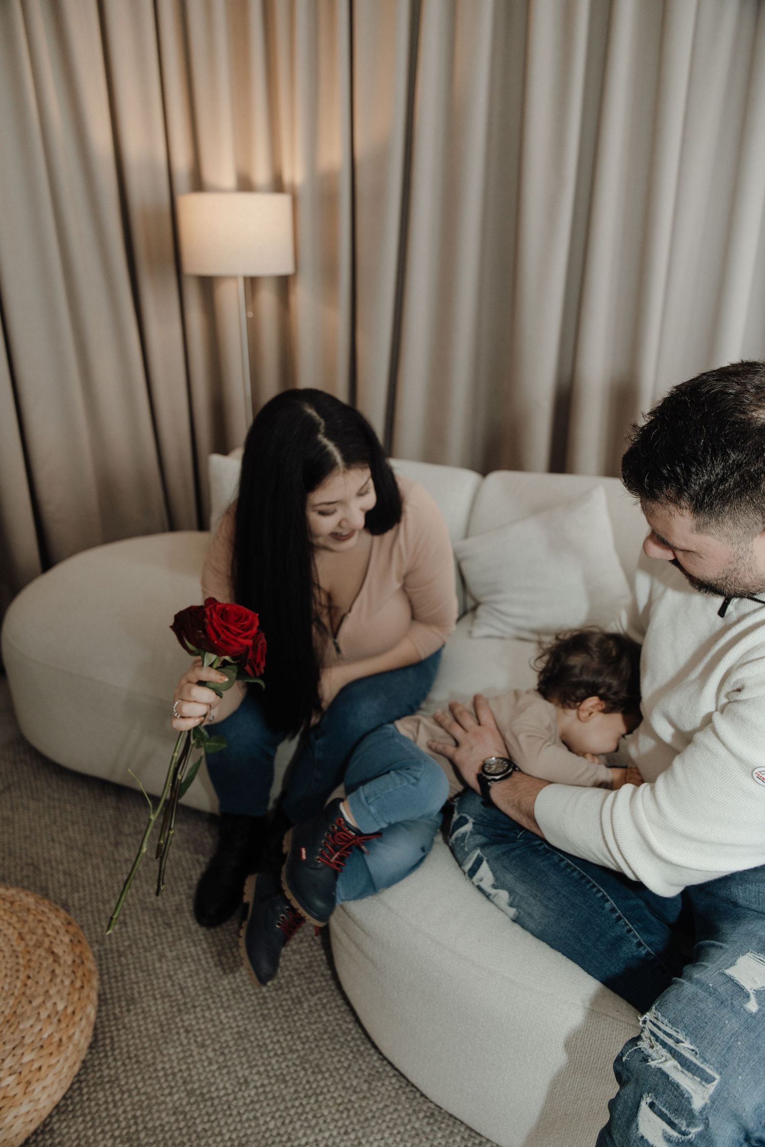 Young couple sits on white sofa, woman holds red roses while man cradles sleeping newborn baby indoors.