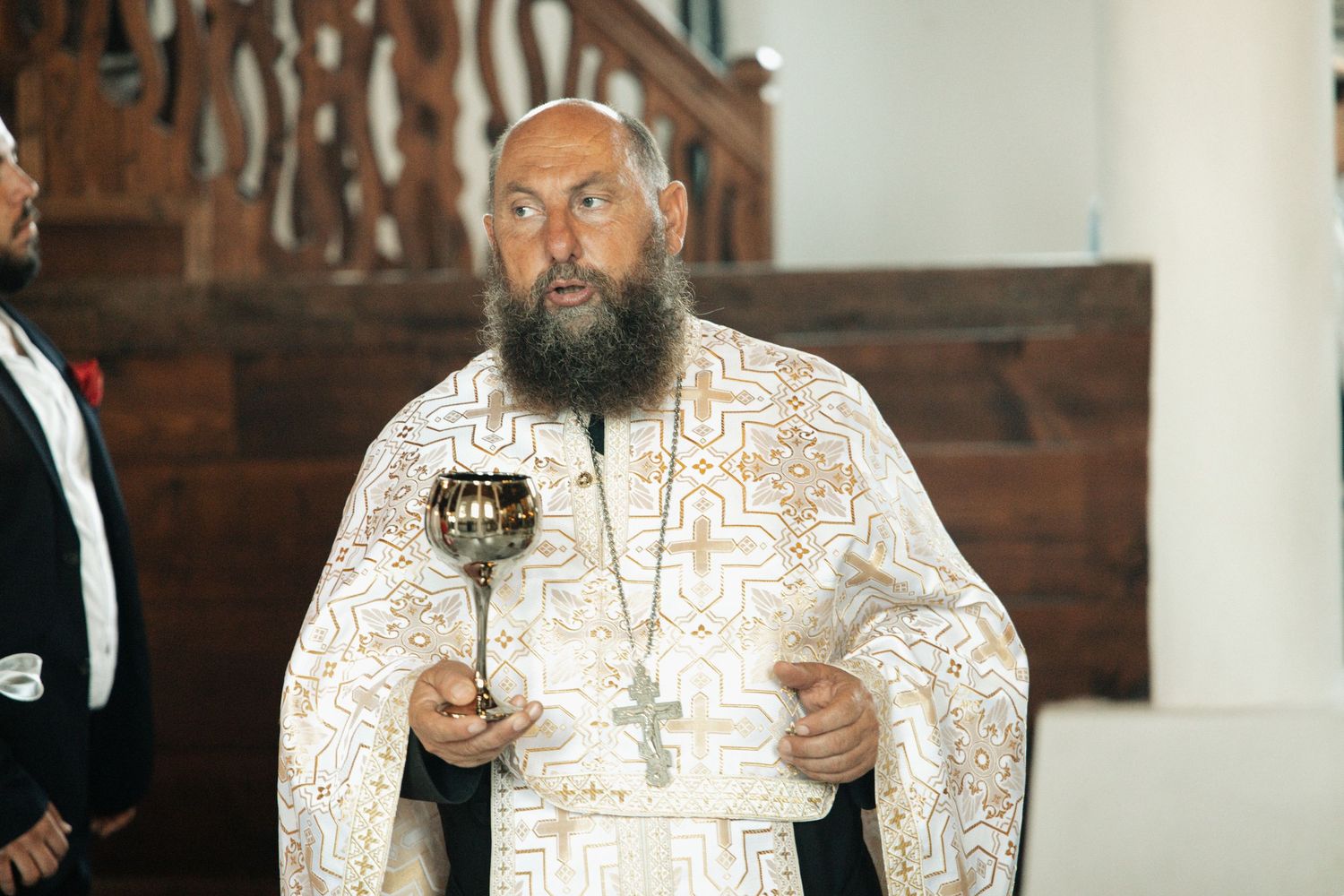 Bearded Orthodox priest in ornate white vestments holding a censer during a religious ceremony.