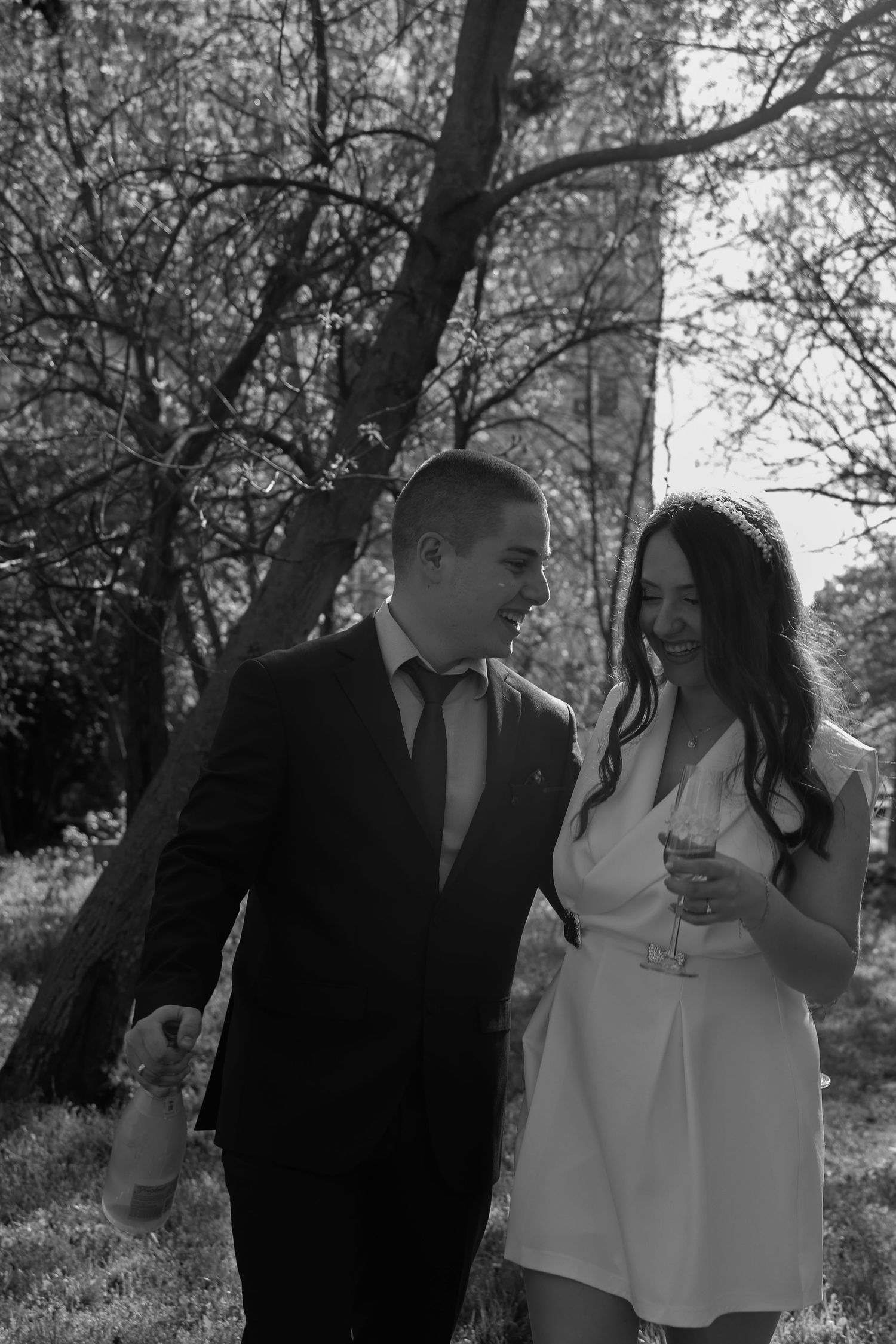 Black and white photo of a smiling couple walking outdoors, man in suit holding bottle, woman in white dress holding champagne glass.