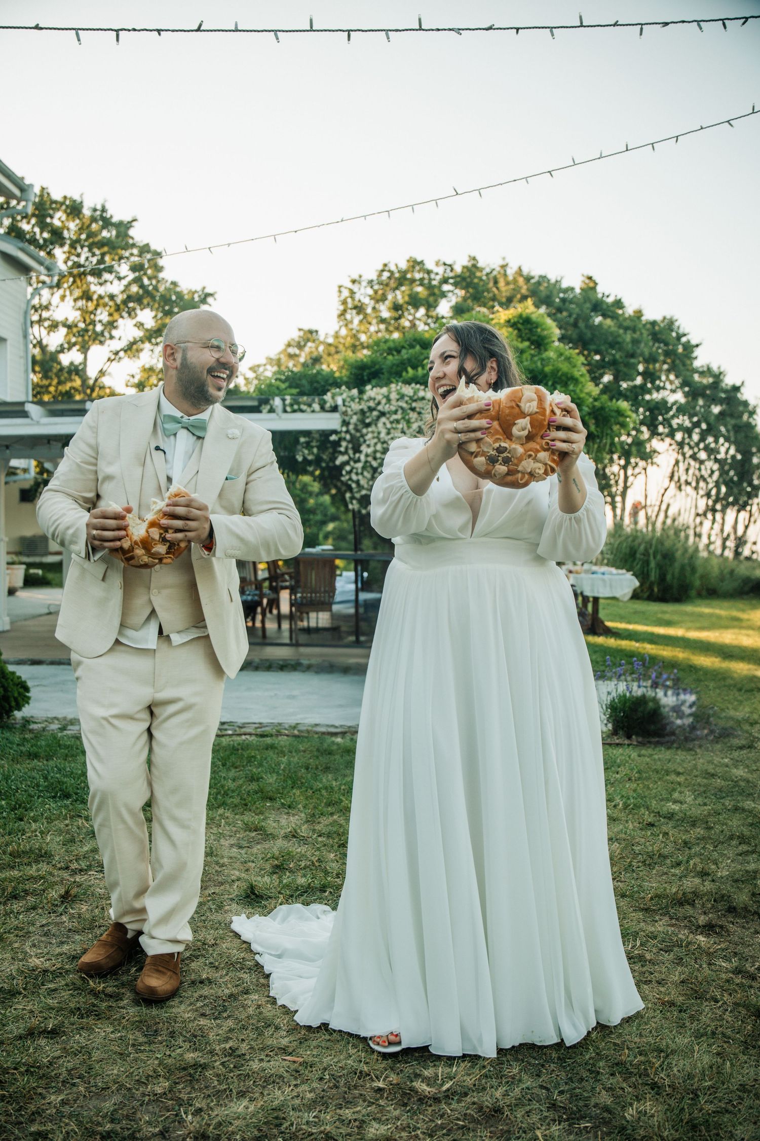 Bride and groom laughing while eating large pretzels outdoors at their wedding reception.