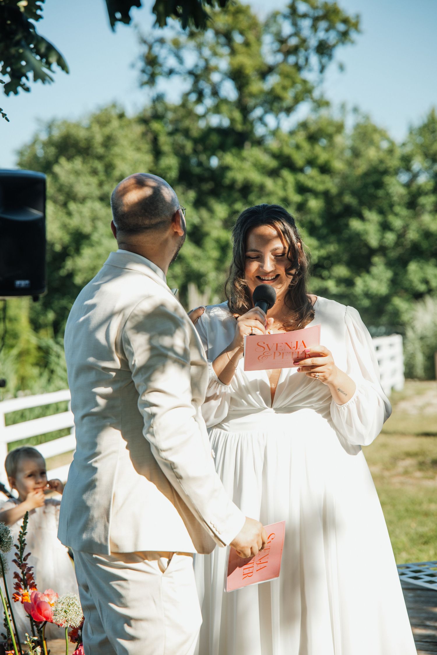 Bride in white gown reading vows from pink card to groom at outdoor wedding ceremony.