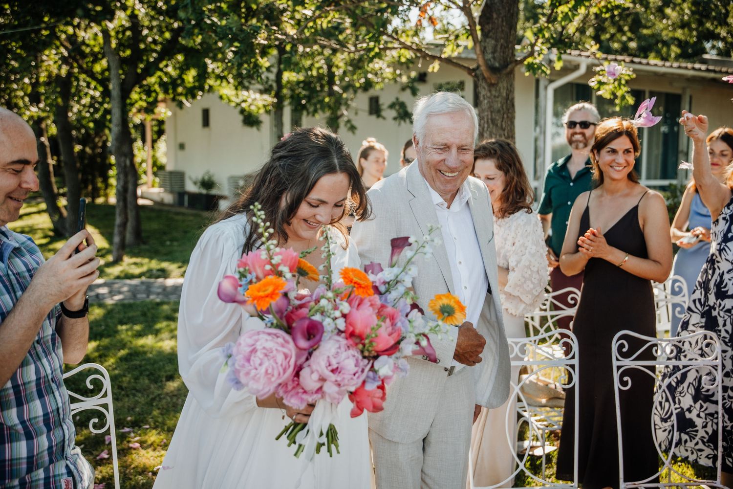 Bride carrying vibrant pink and orange bouquet walks with older gentleman during outdoor wedding ceremony.