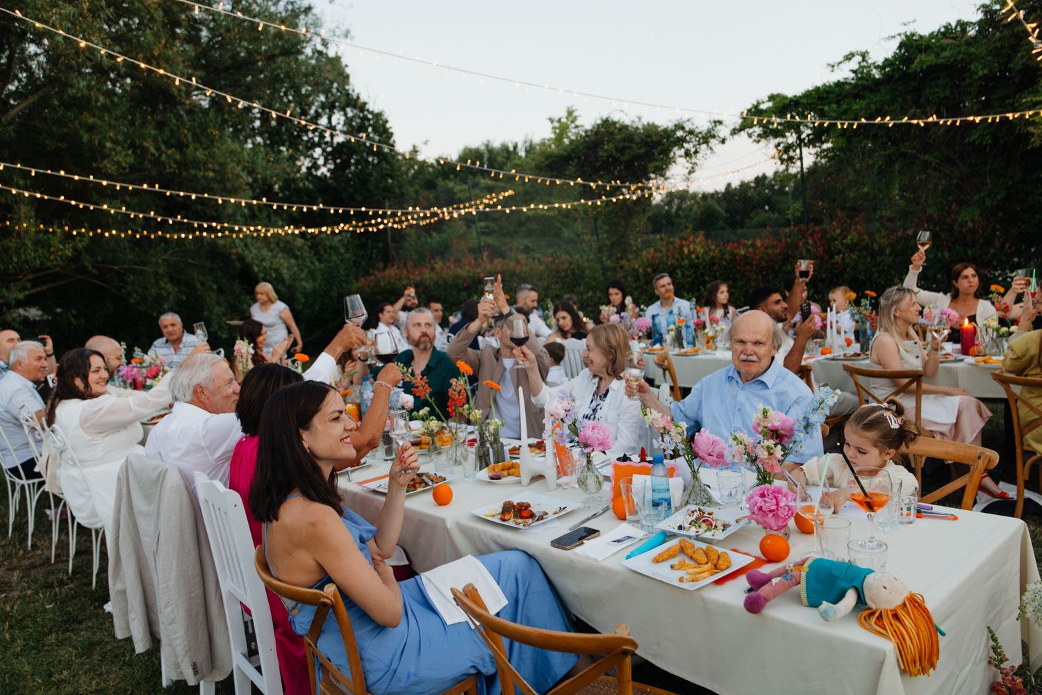 Large outdoor wedding reception with guests raising glasses for a toast under glowing string lights at dusk.