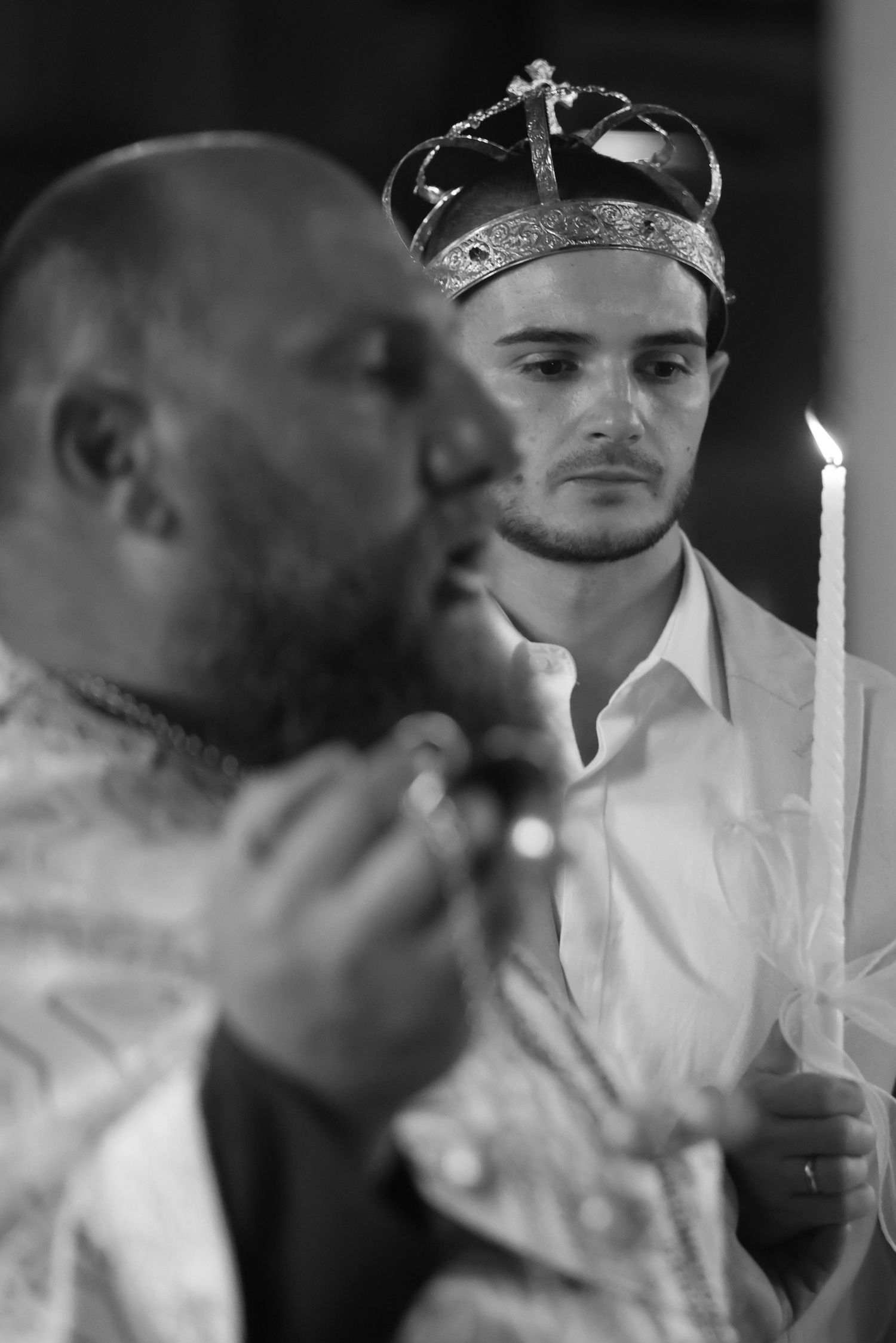 Black and white photo of groom wearing crown holding candle during Orthodox wedding ceremony with priest.