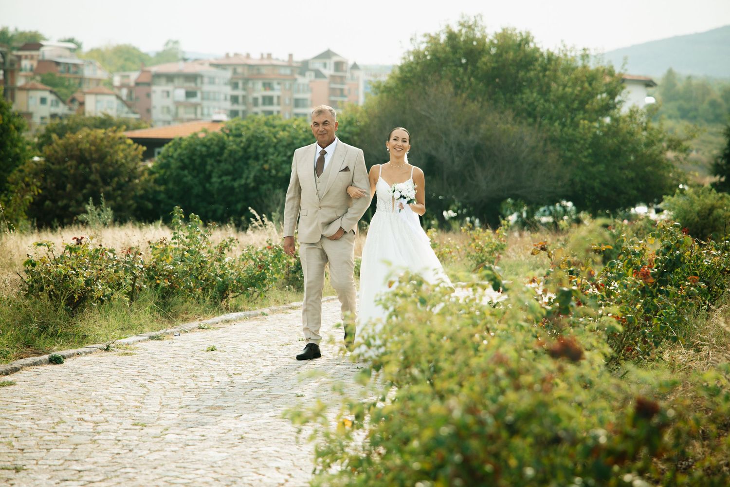 Bride and groom walking hand in hand along a cobblestone path surrounded by lush greenery on their wedding day.