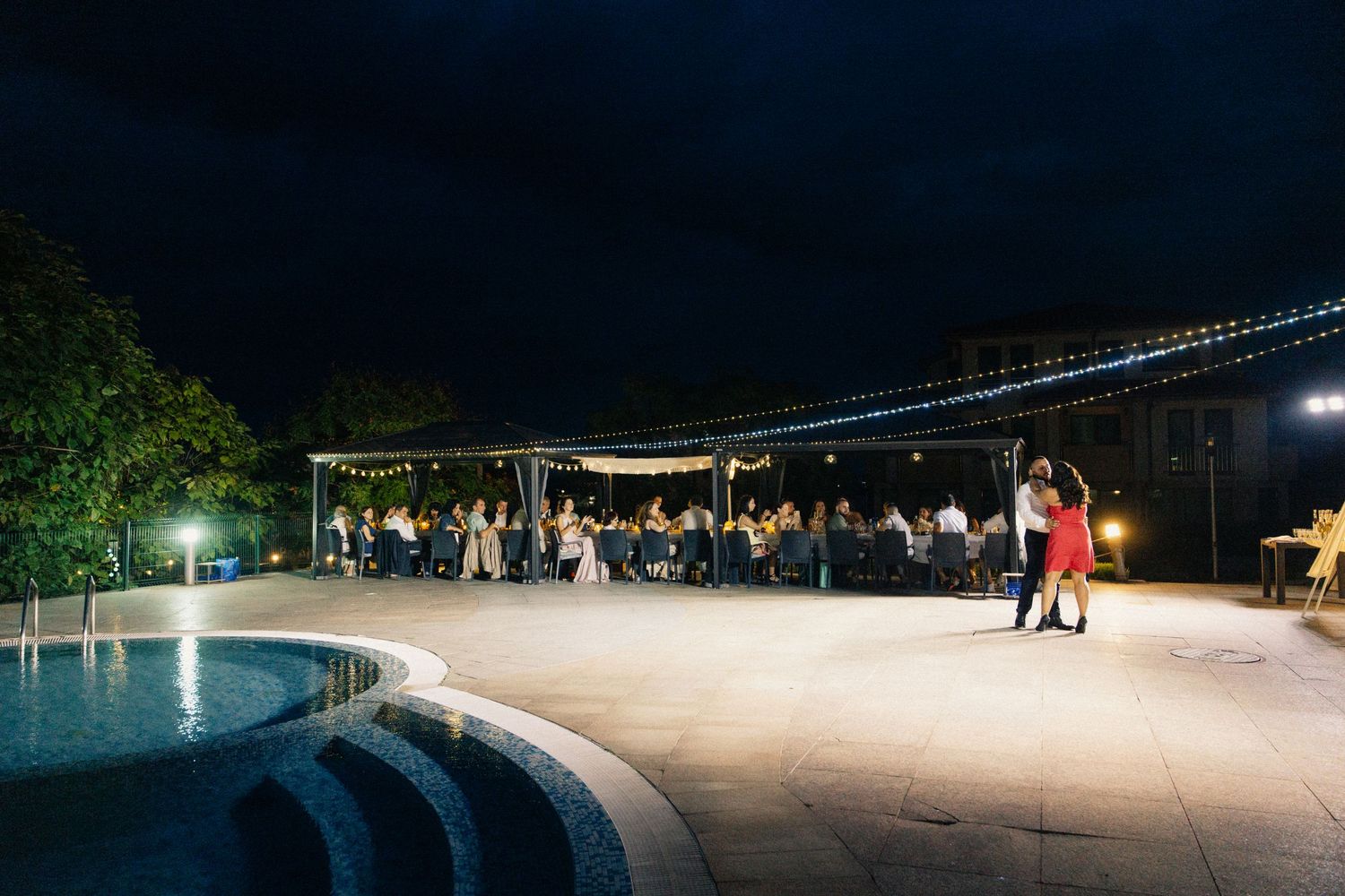 Couple dances on illuminated outdoor patio at night during elegant poolside wedding reception.