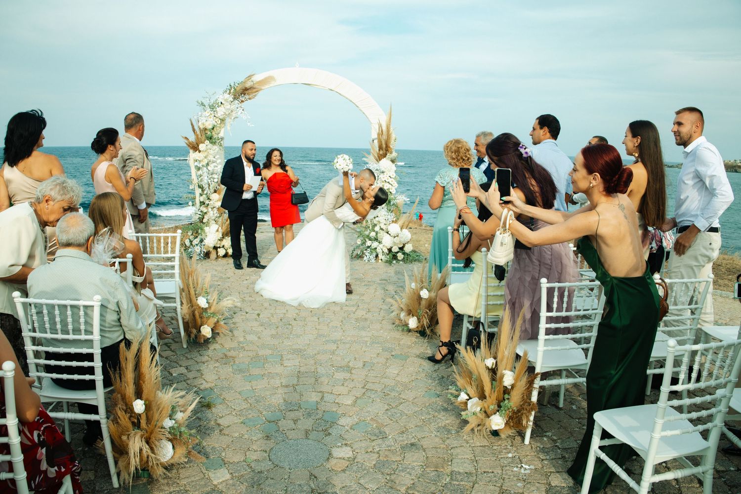Outdoor beach wedding ceremony with guests lining the aisle under a circular floral arch.