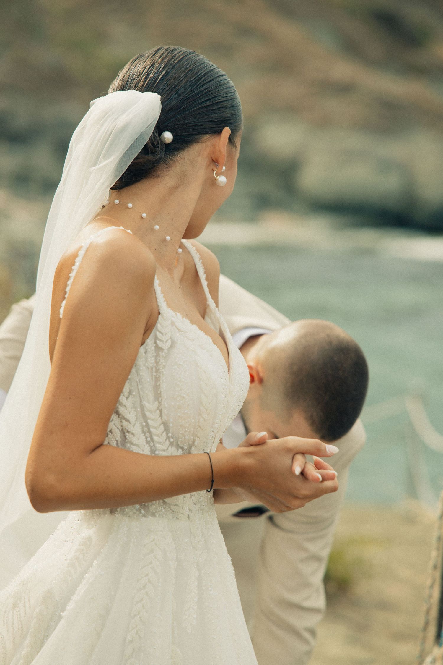 Groom kissing bride's hand at seaside wedding, bride in white lace gown with veil and pearl accessories.