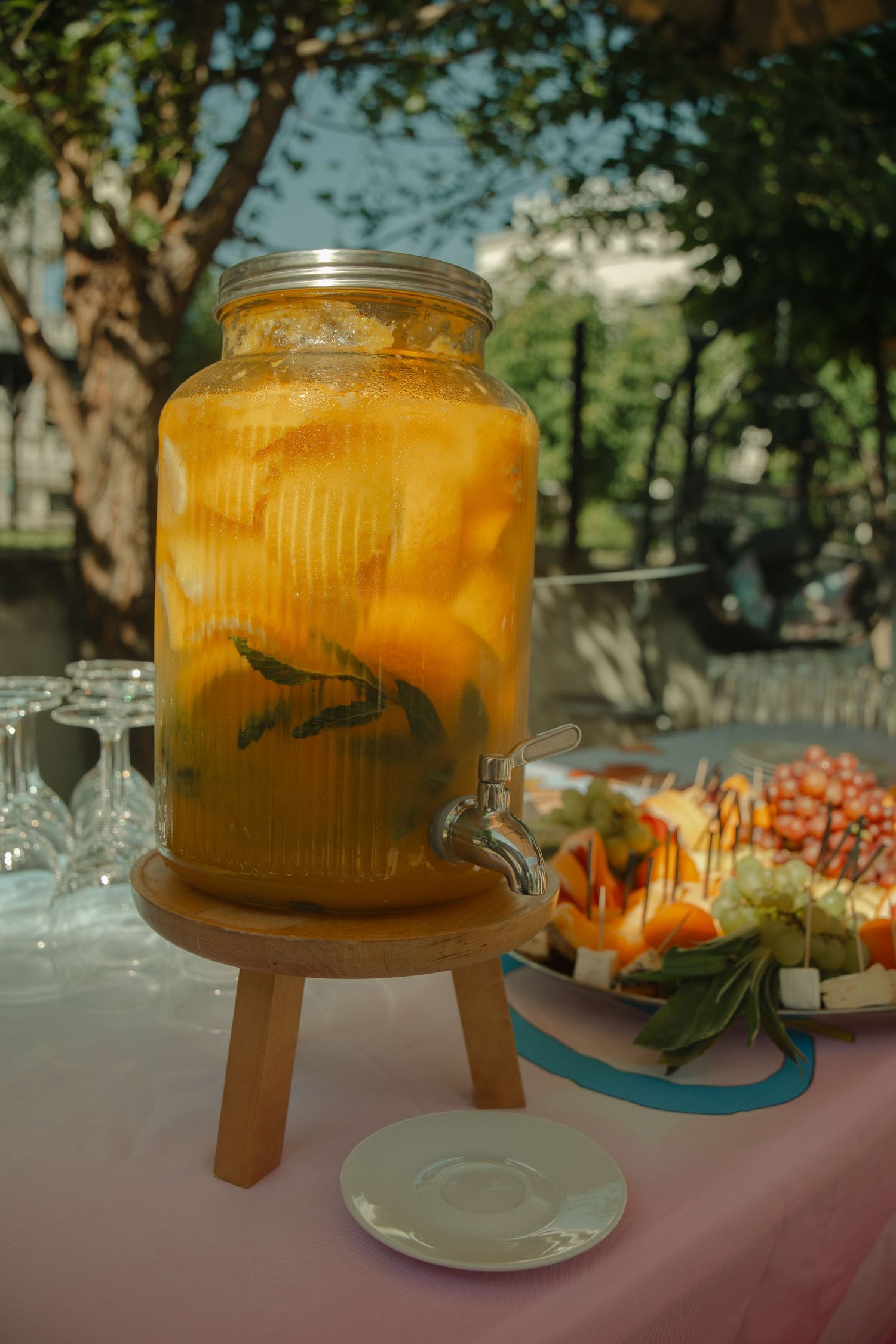 Large glass drink dispenser filled with orange citrus punch on wooden stand at an outdoor party table.