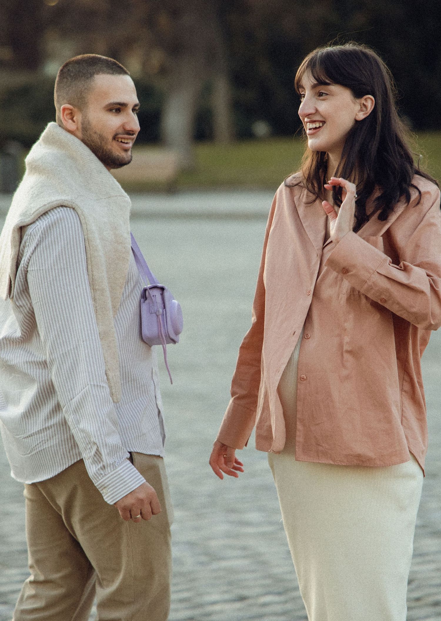 A couple in casual winter outerwear smiling and walking together near a waterfront on an autumn day.
