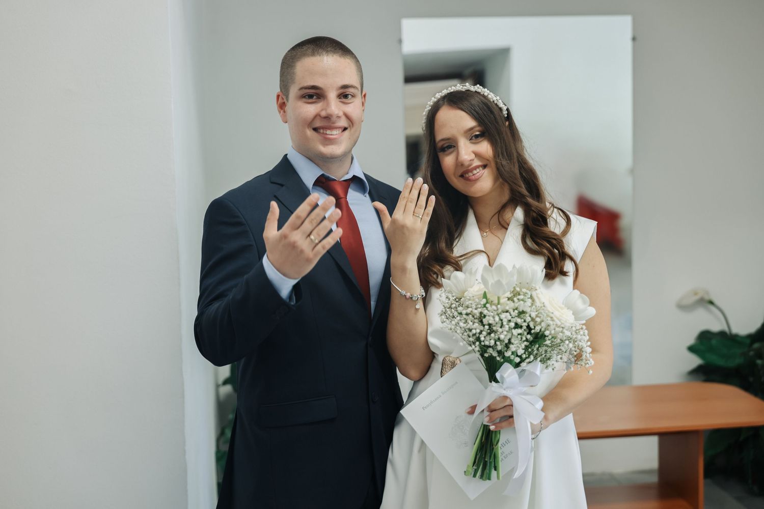 Newlywed couple smiling and showing wedding rings, bride holding white flower bouquet in elegant white dress.