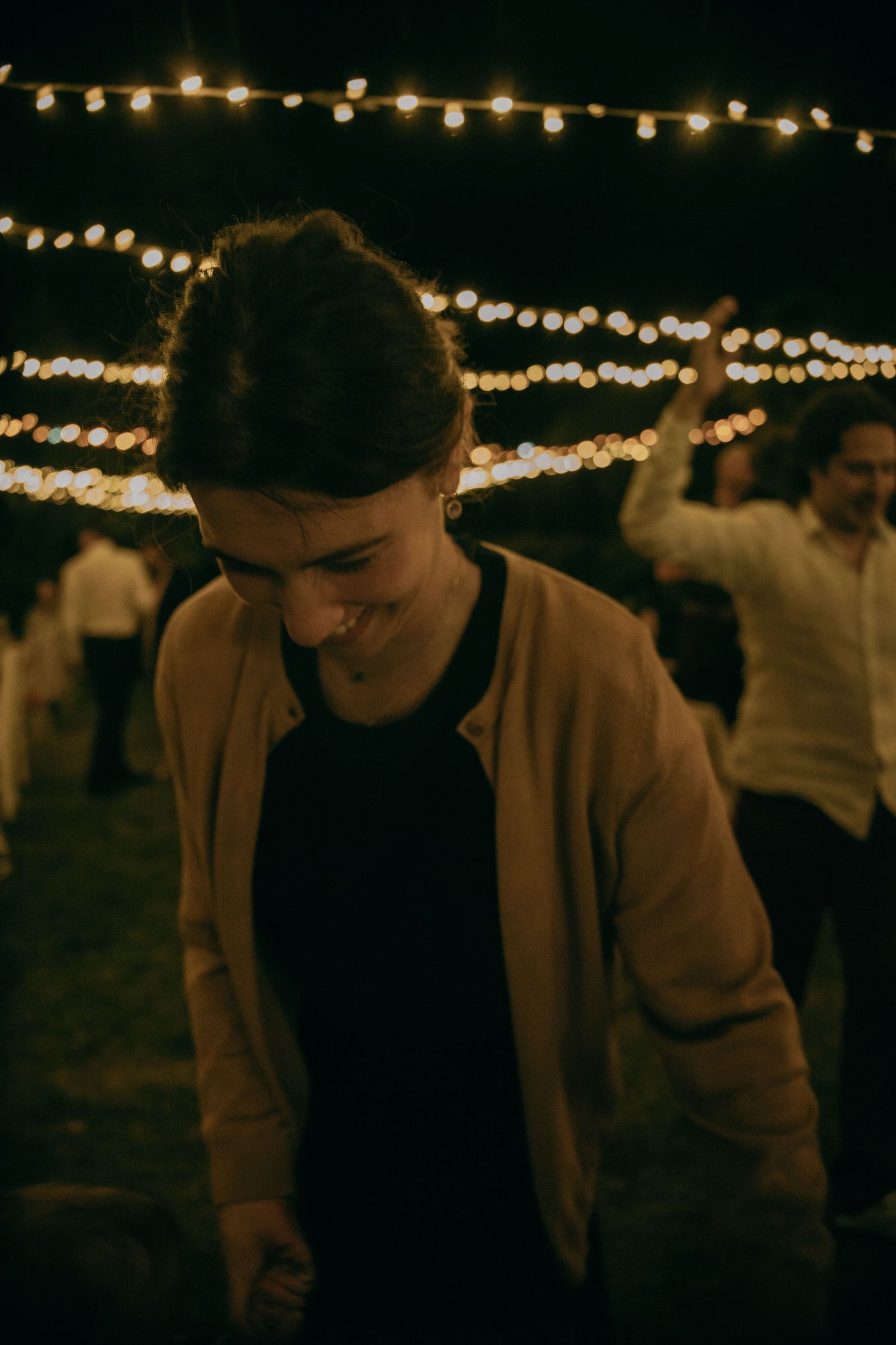 Smiling woman in tan jacket dancing outdoors at night under string lights at a festive evening event.