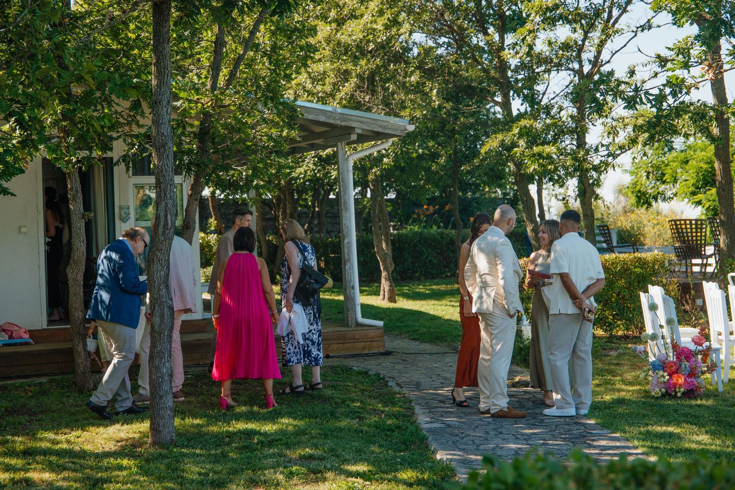Guests mingle on a lush green garden path outside a white cottage during an outdoor wedding event.