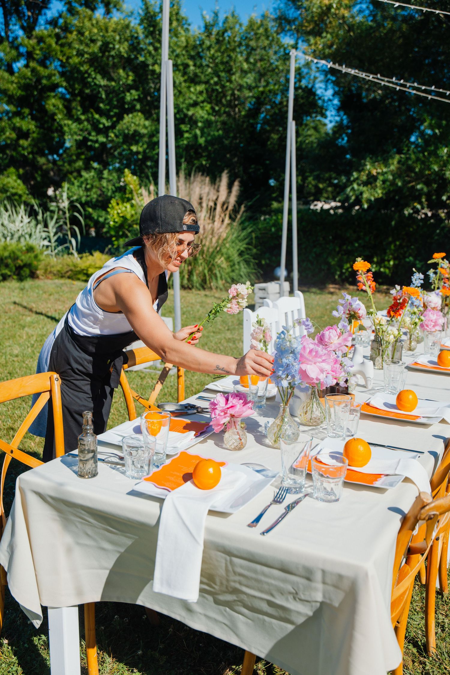 A smiling woman arranges colorful flowers on an outdoor wedding reception table with orange place settings.