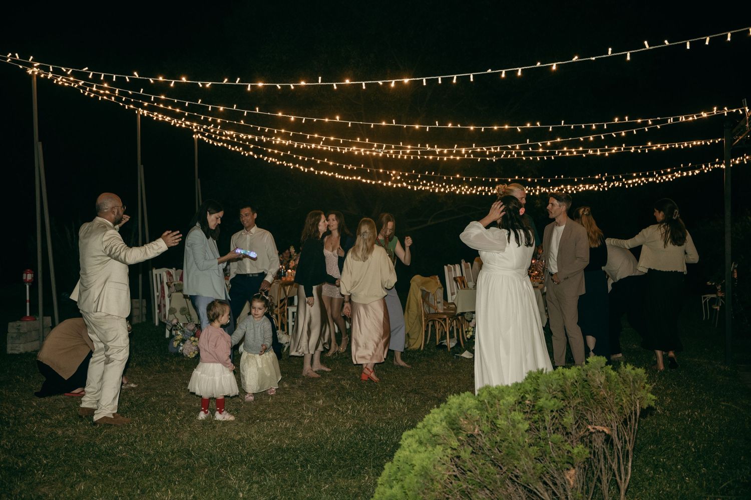 Wedding guests dance under canopy of warm string lights at outdoor evening celebration.