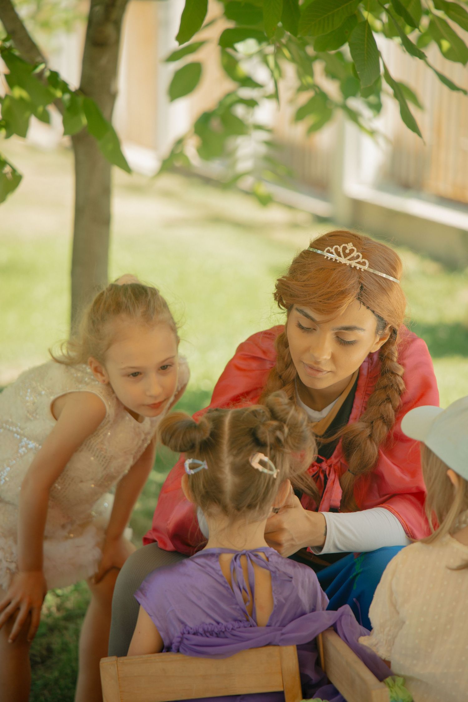 Princess character in red costume braiding young girl's hair at outdoor children's party.