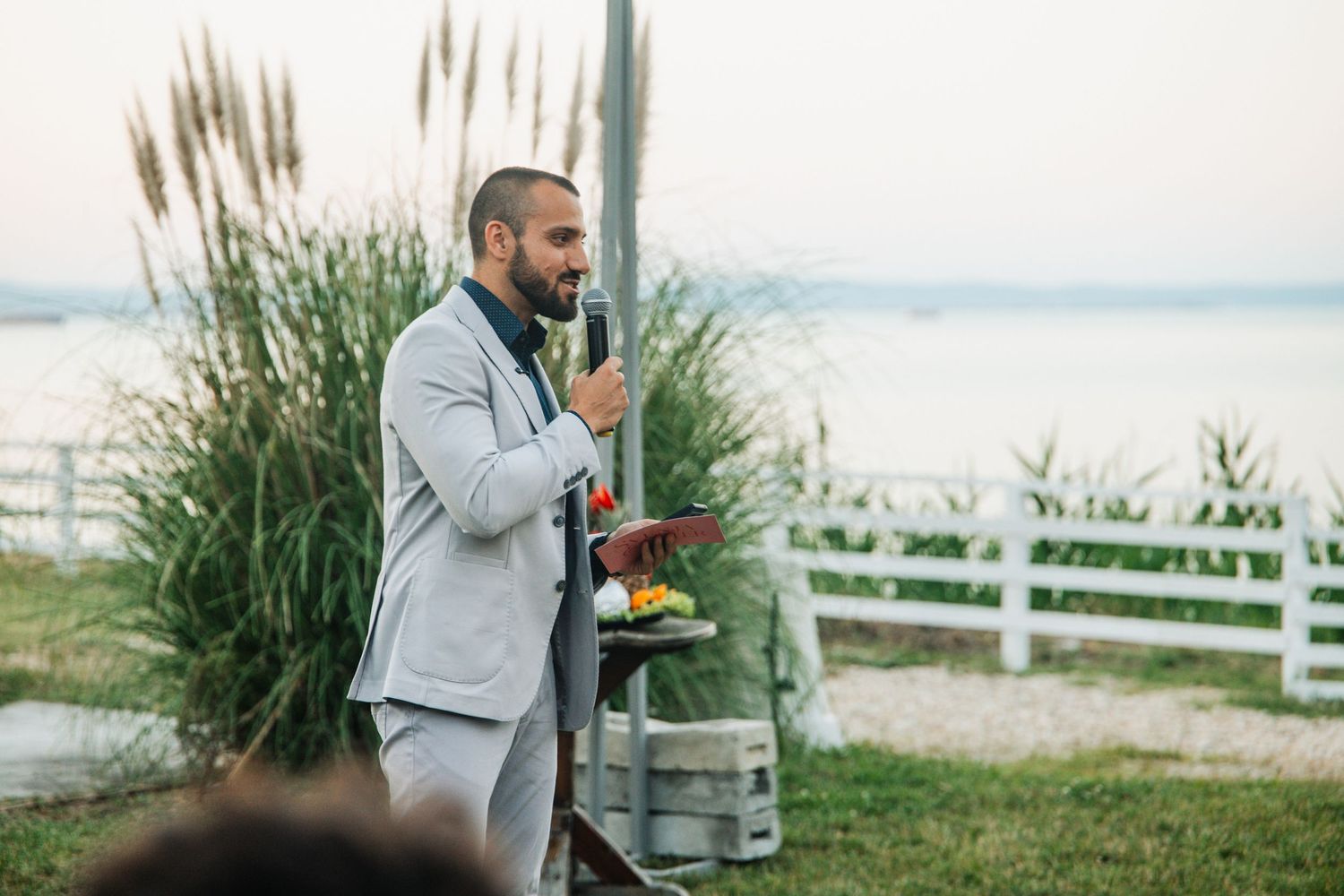 Groom in grey suit giving speech at outdoor lakeside wedding ceremony near white fence at sunset.