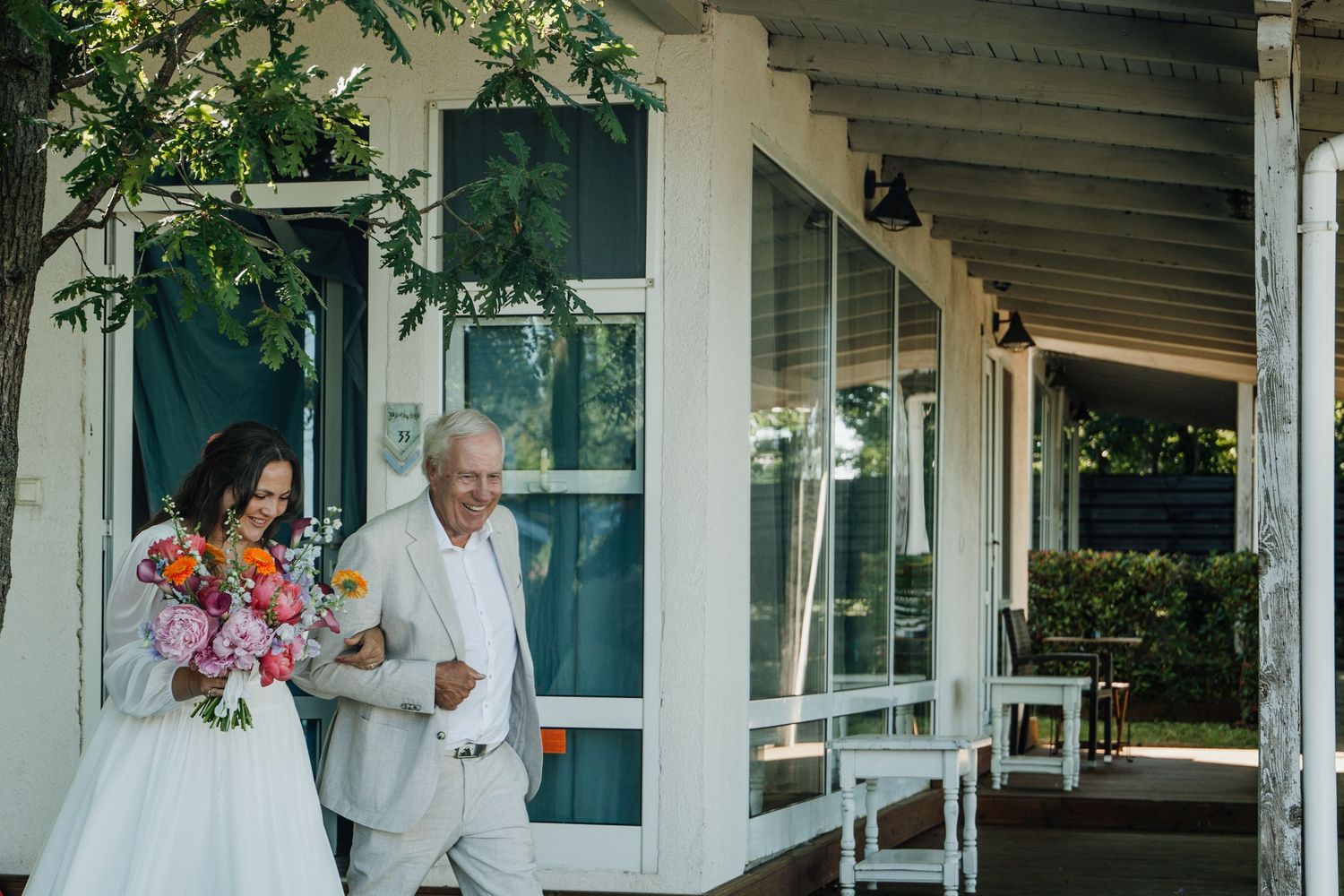 Bride in white gown holding colorful bouquet walks with older gentleman along venue porch before ceremony.