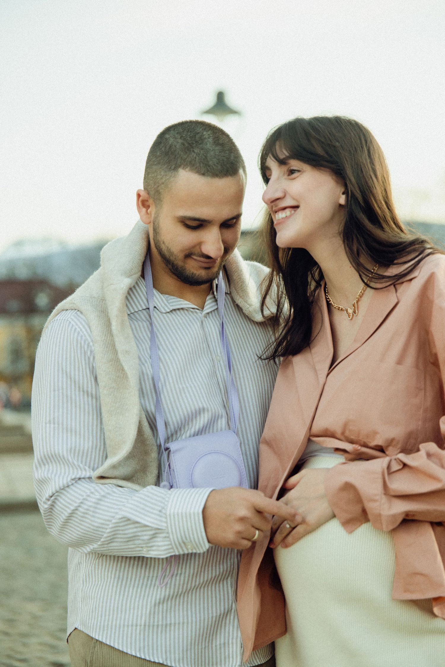 A couple shares a tender moment outdoors, with one wearing a striped shirt and camera while the other wears a pink blazer.