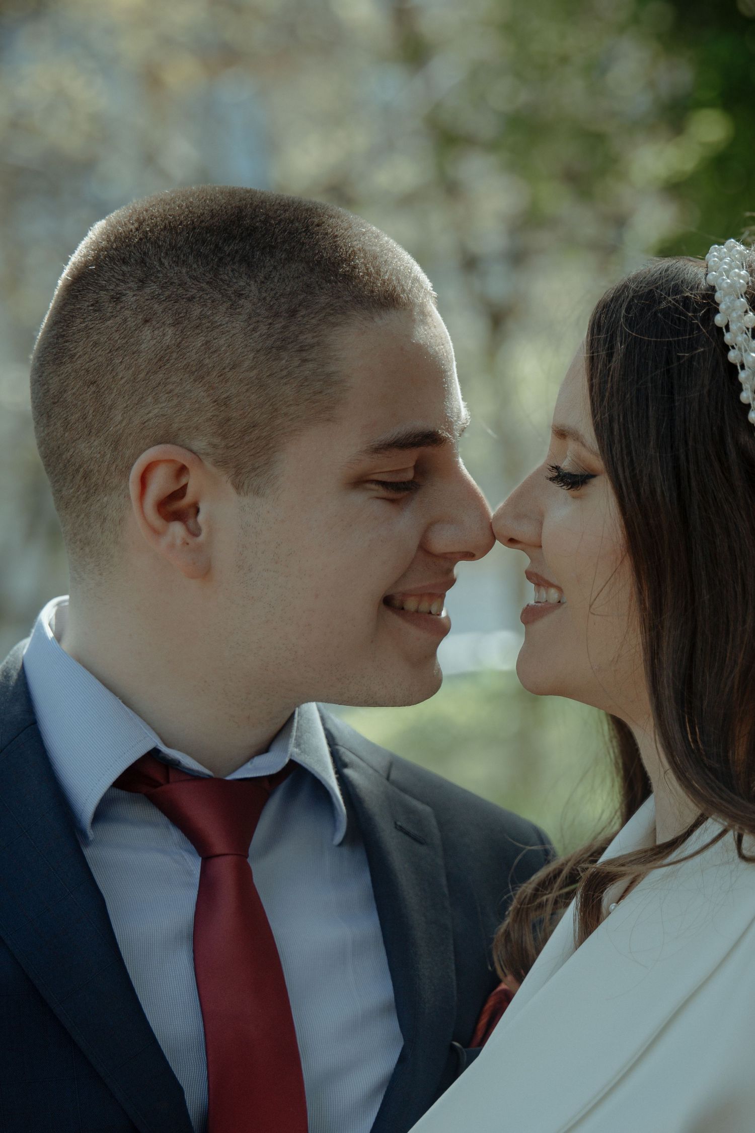 A smiling bride and groom share a romantic nose-to-nose moment, with the groom in a suit and red tie.