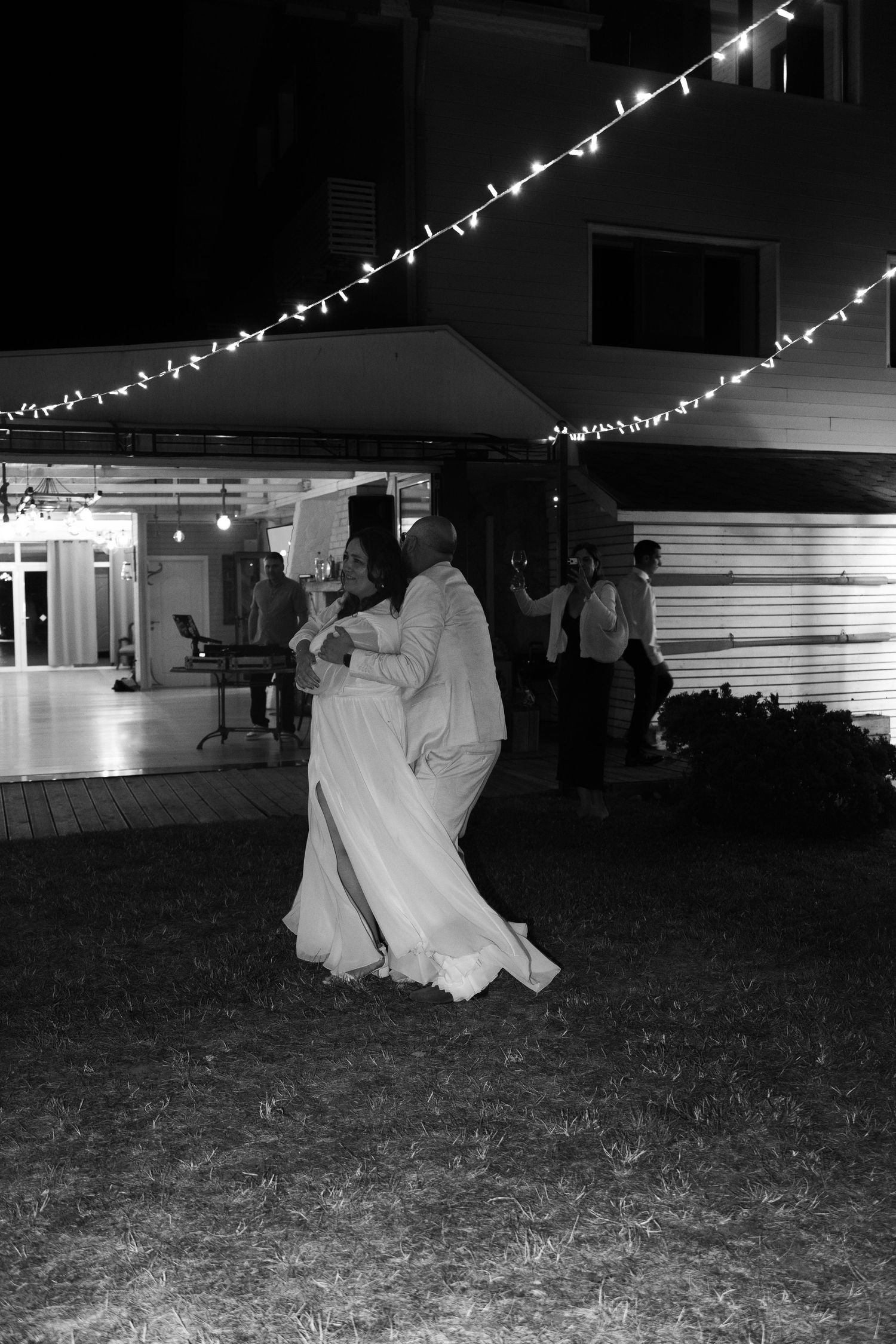 Black and white photo of a couple sharing their first dance outdoors at a wedding reception with string lights.